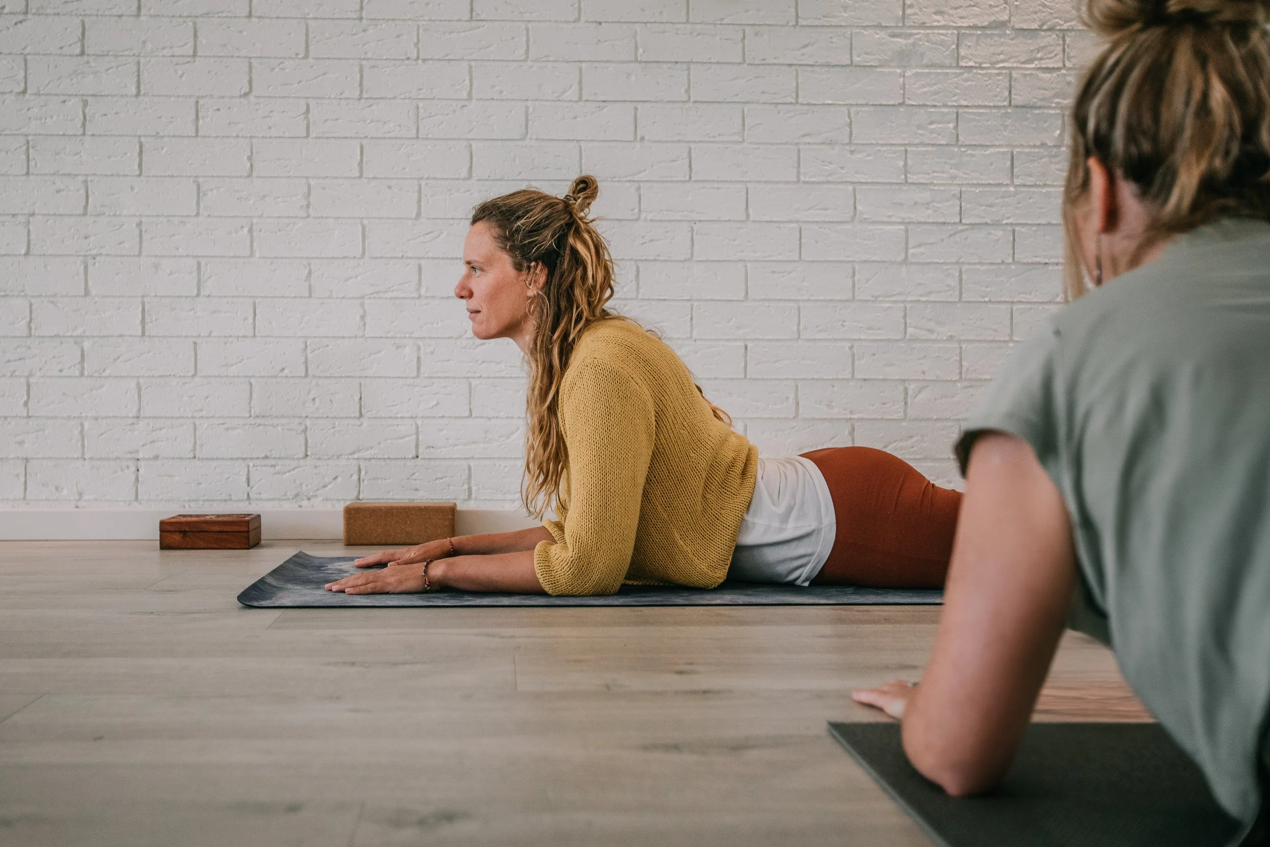 Two women practicing yoga indoors on mats, with one woman in a yellow sweater and brown pants doing the Cobra pose, while the other woman is in the foreground with her arm bent in a similar pose.