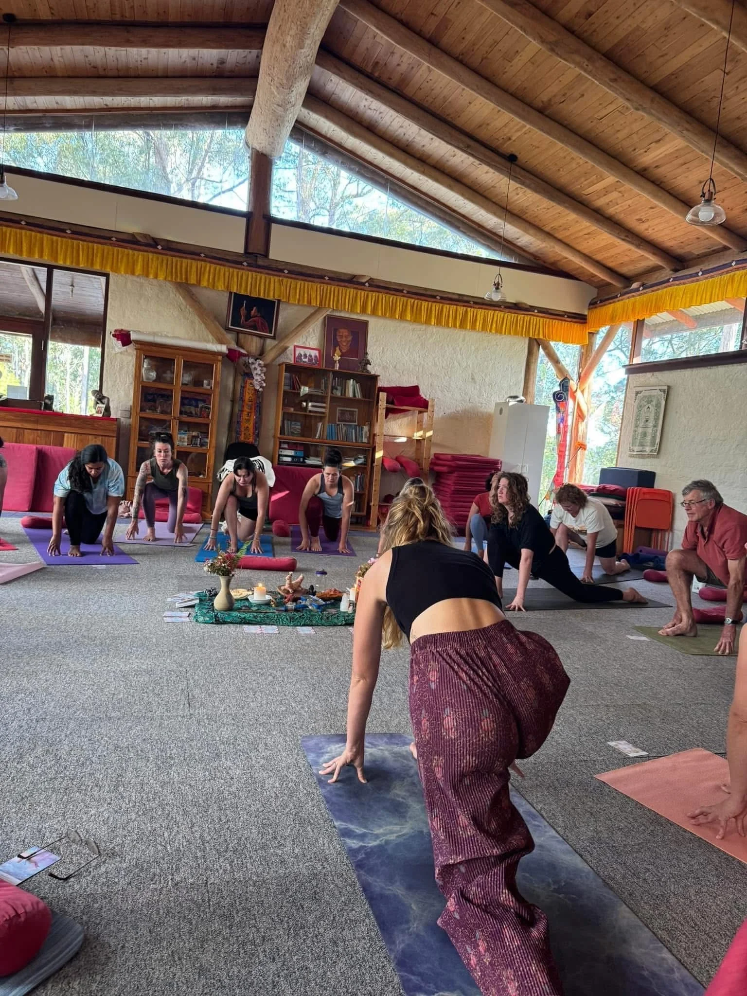 A group of people participating in a yoga class in an indoor studio with wooden ceiling beams, large windows, and religious decorations, practicing stretches on yoga mats.