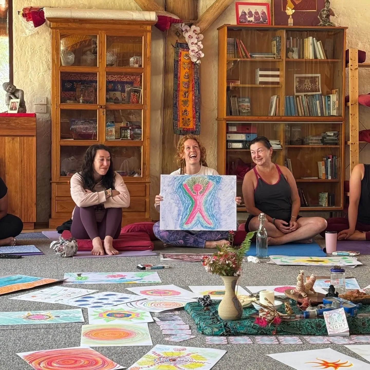 Group of women seated on the floor during a workshop or class, with colorful drawings and paintings spread out in front of them; one woman in the middle holding up a drawing of a colorful, abstract human figure with raised arms, and laughing. The roo