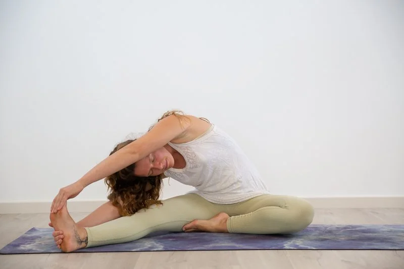 Woman practicing yoga seated on a mat, stretching with one arm over her head and holding her foot.