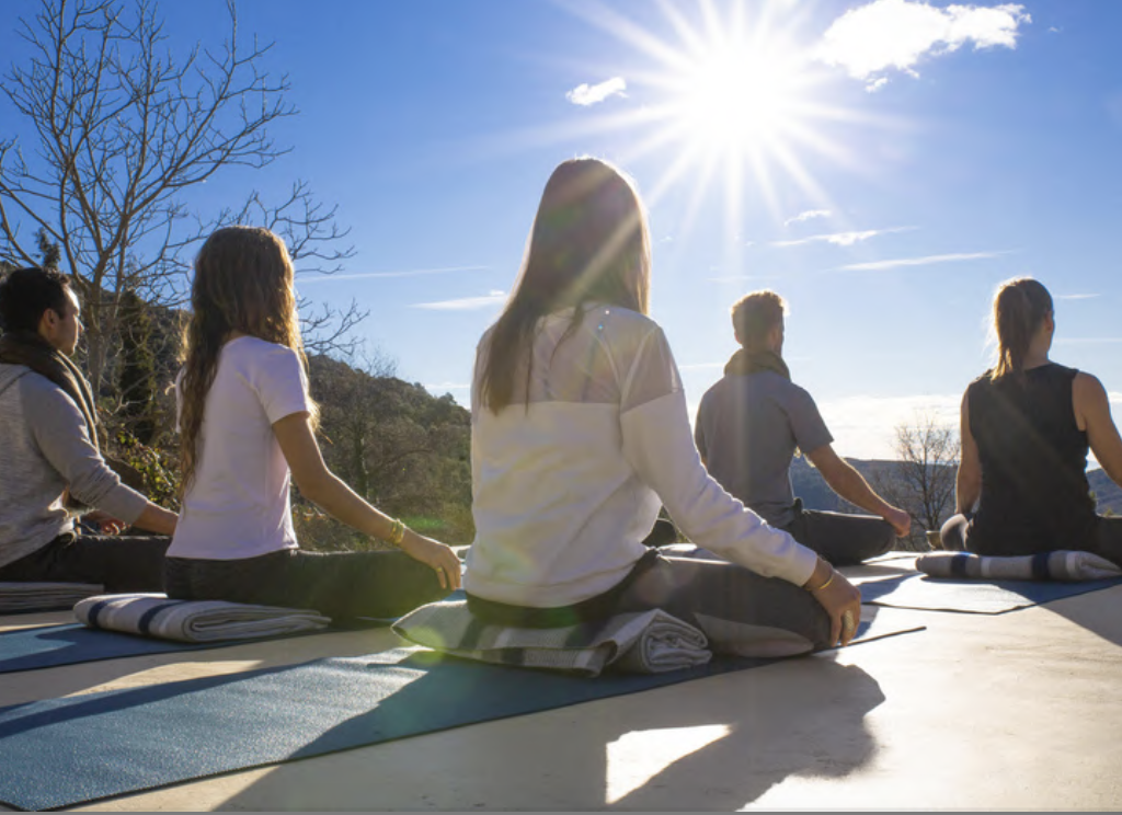 Woman with long blonde hair wearing a purple patterned dress and sandals walking along a narrow tiled pathway in a courtyard with intricate Moroccan-style architecture and lush garden.