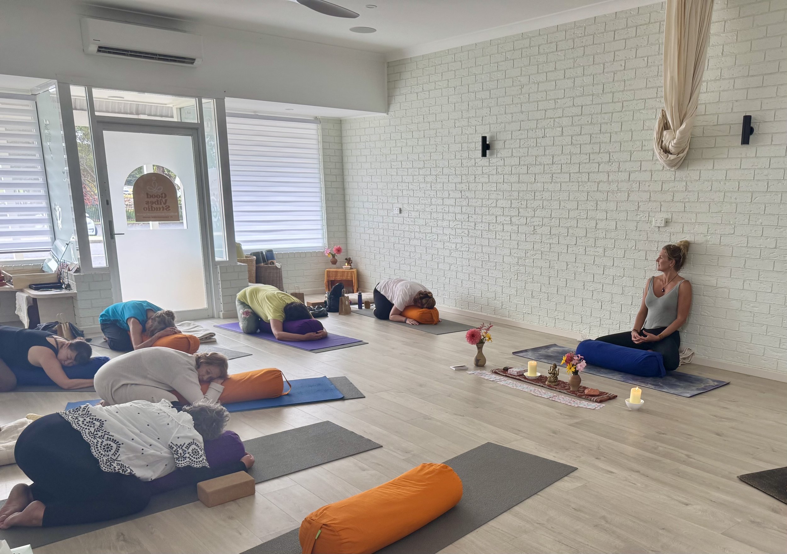 A yoga class with six women practicing poses on mats in a bright, airy studio with white brick walls, large windows with blinds, and floral decorations.