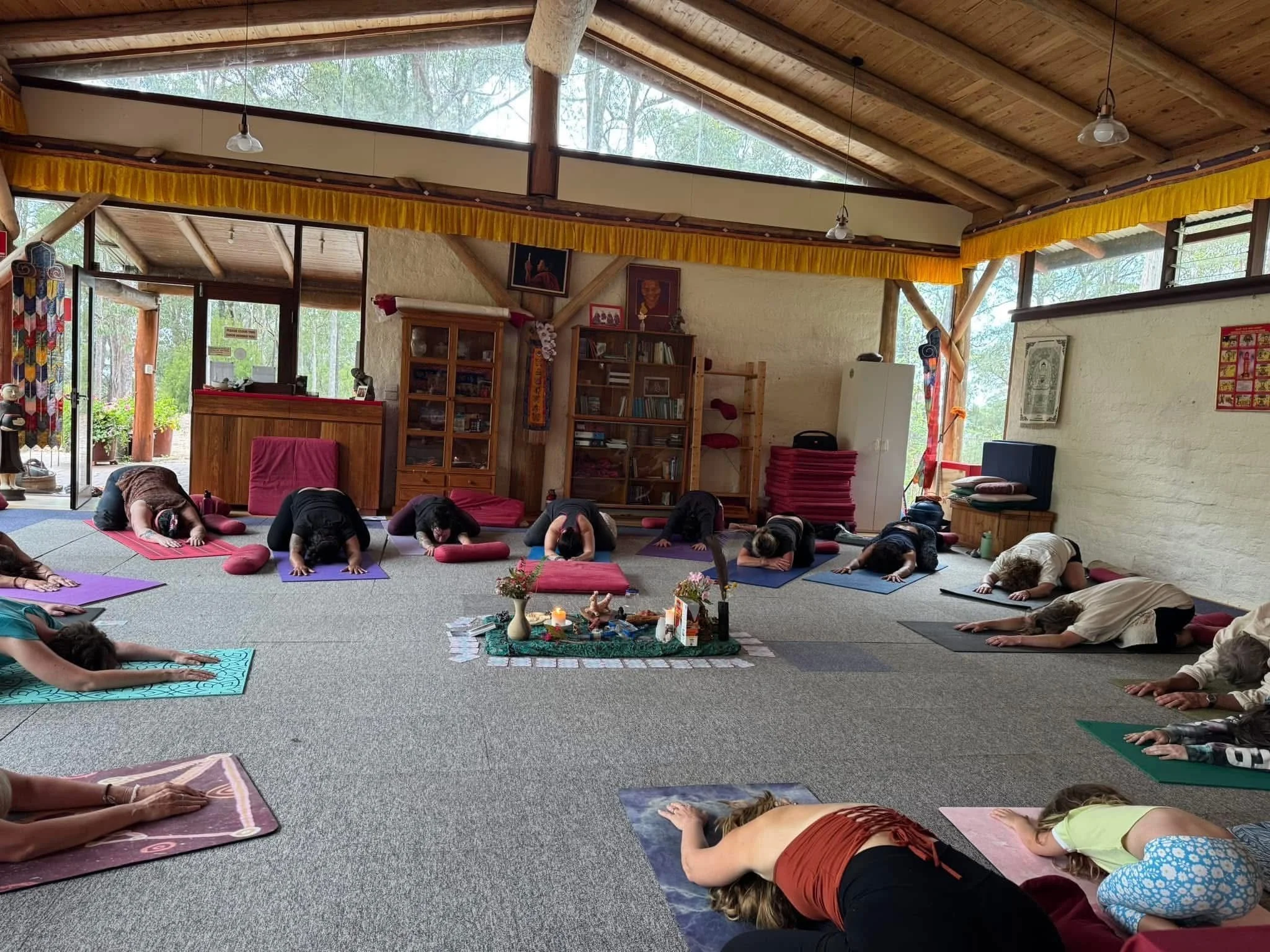 A group of people participating in a yoga class in a bright, spacious room with large windows, wooden ceiling beams, and yoga mats on the floor, some with red cushions, practicing a child's pose.