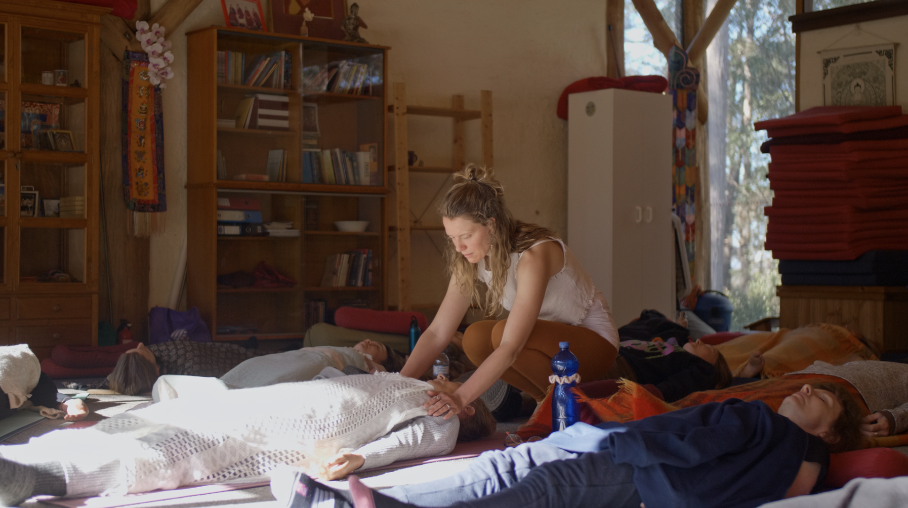 A woman is giving a guided meditation to a group of people lying on the floor in a cozy room with bookshelves and large windows.