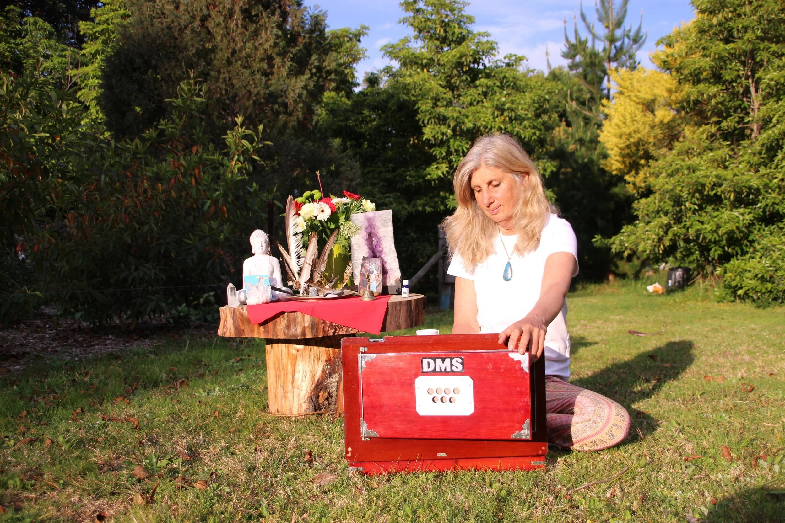 A woman with long blonde hair sitting outdoors on grass, reaching into a red box labeled 'DMS'. Behind her is a wooden table with religious and spiritual items, including a small Buddha statue, flowers, and crystals, with trees and blue sky in the background.