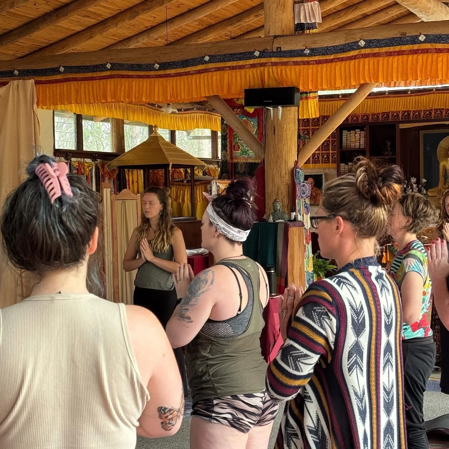 Group of women participating in a yoga or meditation class inside a decorated wooden room with orange curtains and cultural artwork.