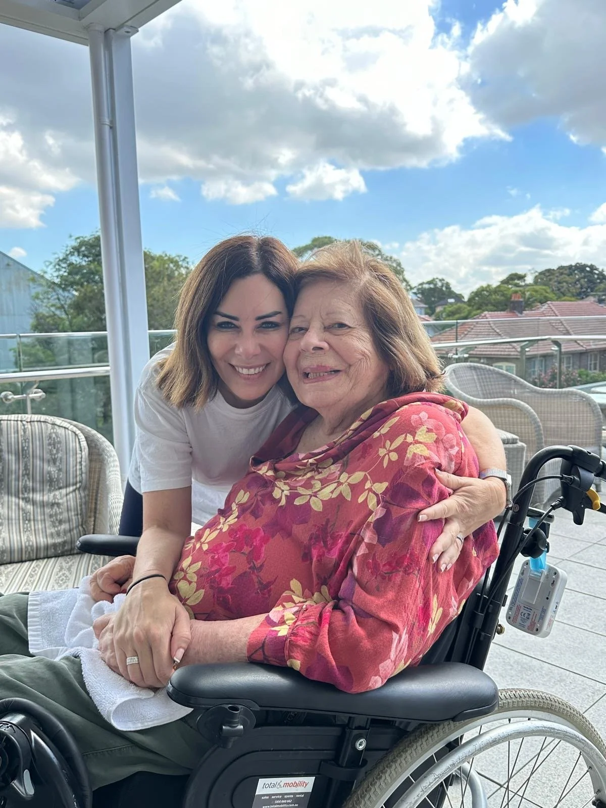 A young woman and an elderly woman sitting together outdoors on a patio, smiling and hugging, with a clear sky and trees in the background.