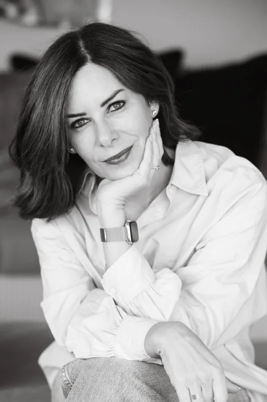 Black and white portrait of a woman with shoulder-length dark hair, wearing a white button-down shirt, resting her head on her hand, and looking at the camera with a slight smile.