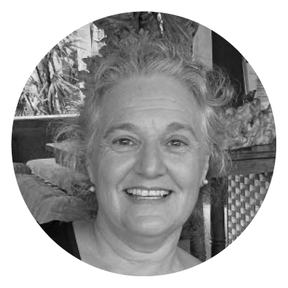 Black and white photo of a smiling woman with curly hair, sitting in a room with plants and a wicker chair in the background.