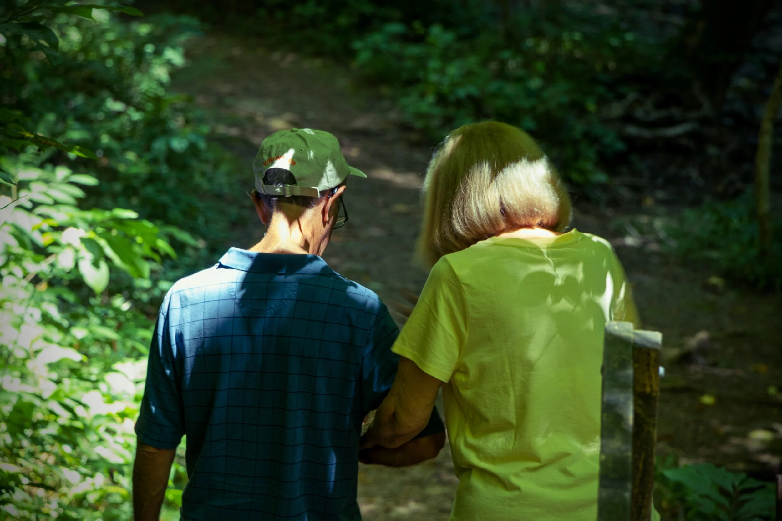 Two elderly people, one man wearing a green cap and a blue checkered shirt and a woman wearing a yellow t-shirt, walk together on a forest trail, holding hands or assisting each other.