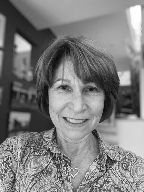 A woman with short hair smiling, wearing a patterned shirt and a heart-shaped necklace in an indoor setting.