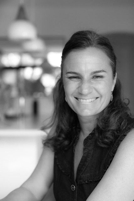 Black and white photo of a woman smiling with long hair, wearing a sleeveless top, indoors in a well-lit space.
