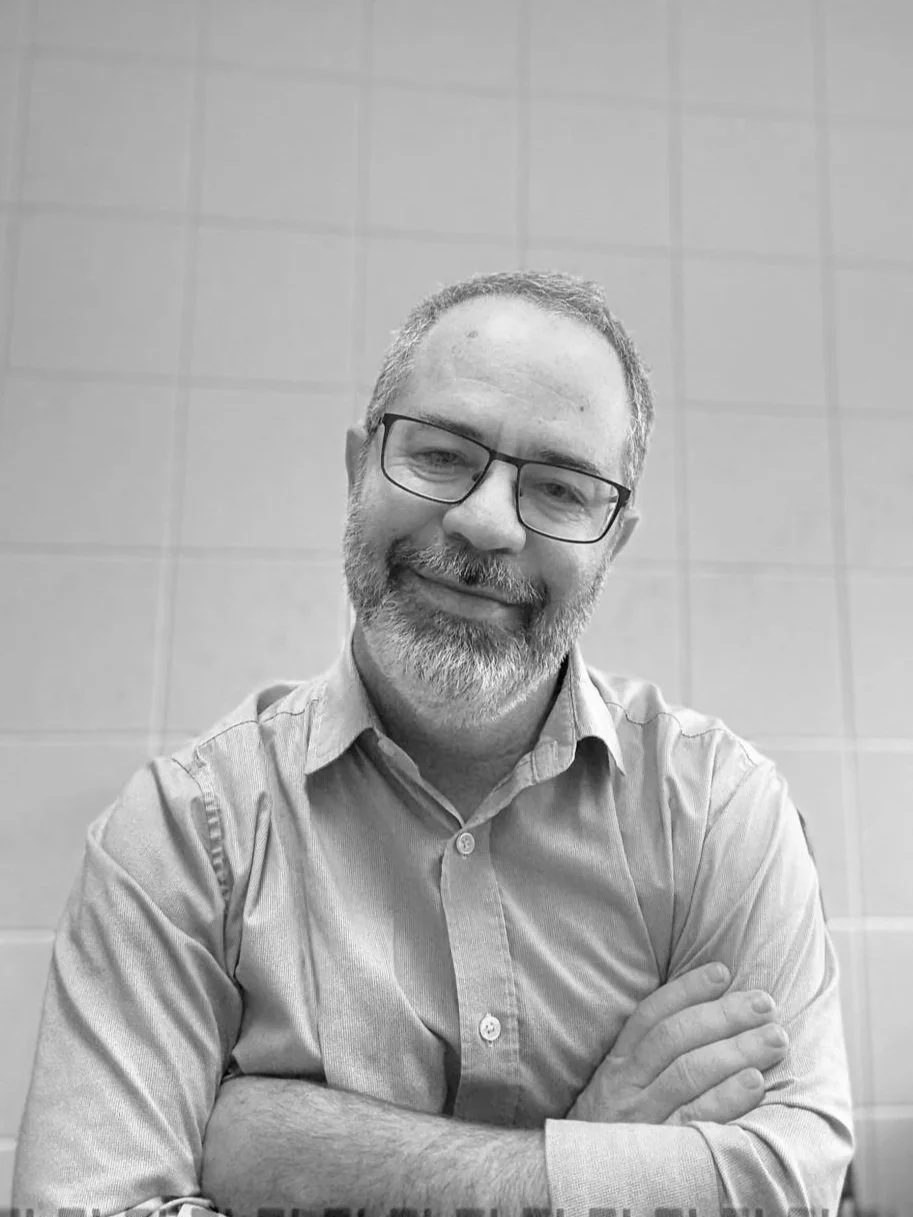 A black-and-white photo of a smiling middle-aged man with glasses, a beard, and short hair, wearing a button-up shirt, sitting with his arms crossed in front of a tiled wall.