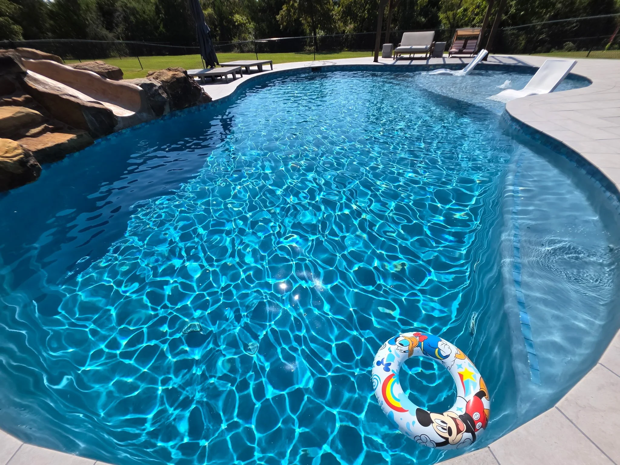 Outdoor swimming pool with rock waterfall, slide, and lounge chairs on a sunny day.