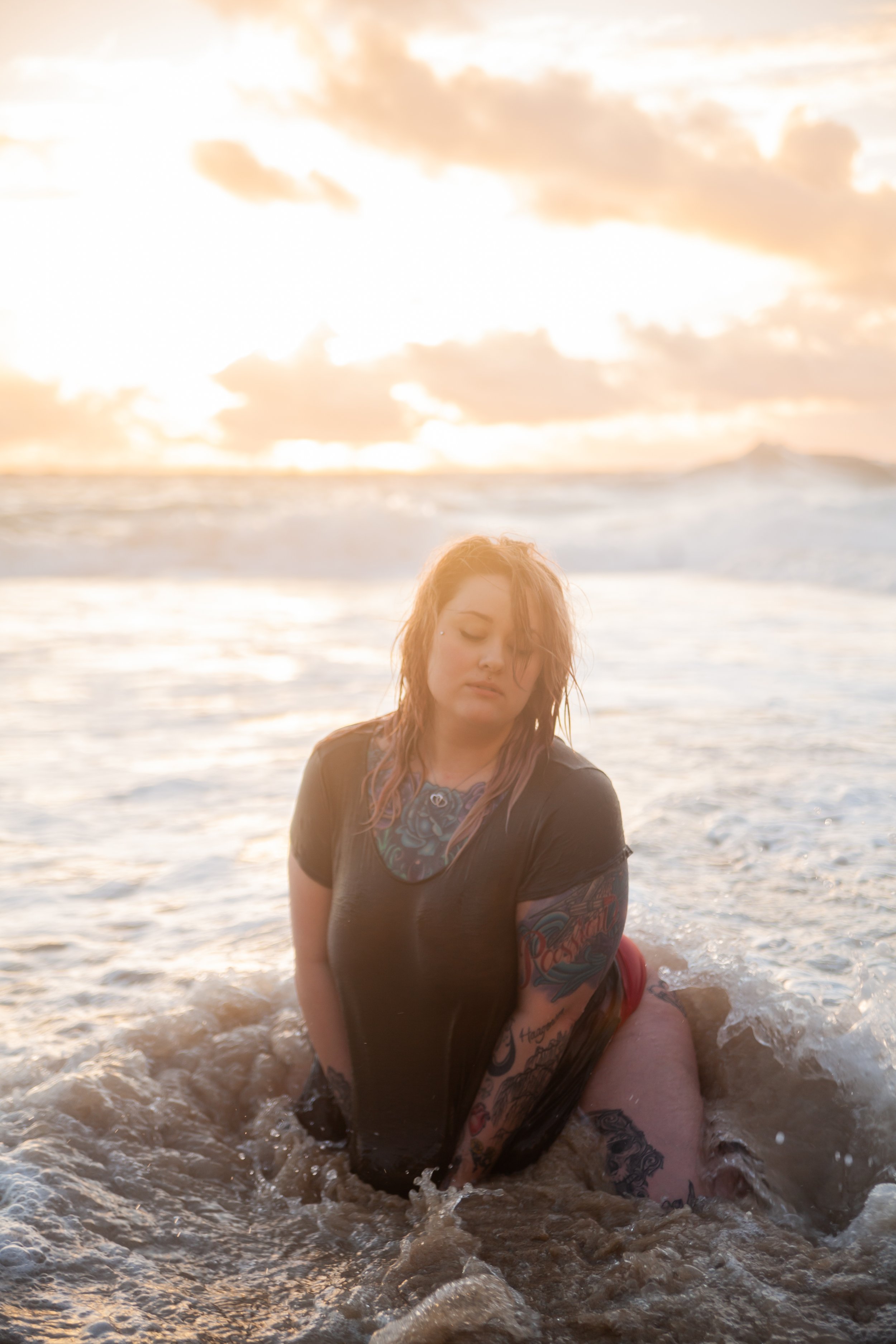 A young woman with dyed hair and tattoos on her arms and chest, sitting in the shallow water at the beach during sunset, with her eyes closed and a contemplative expression.