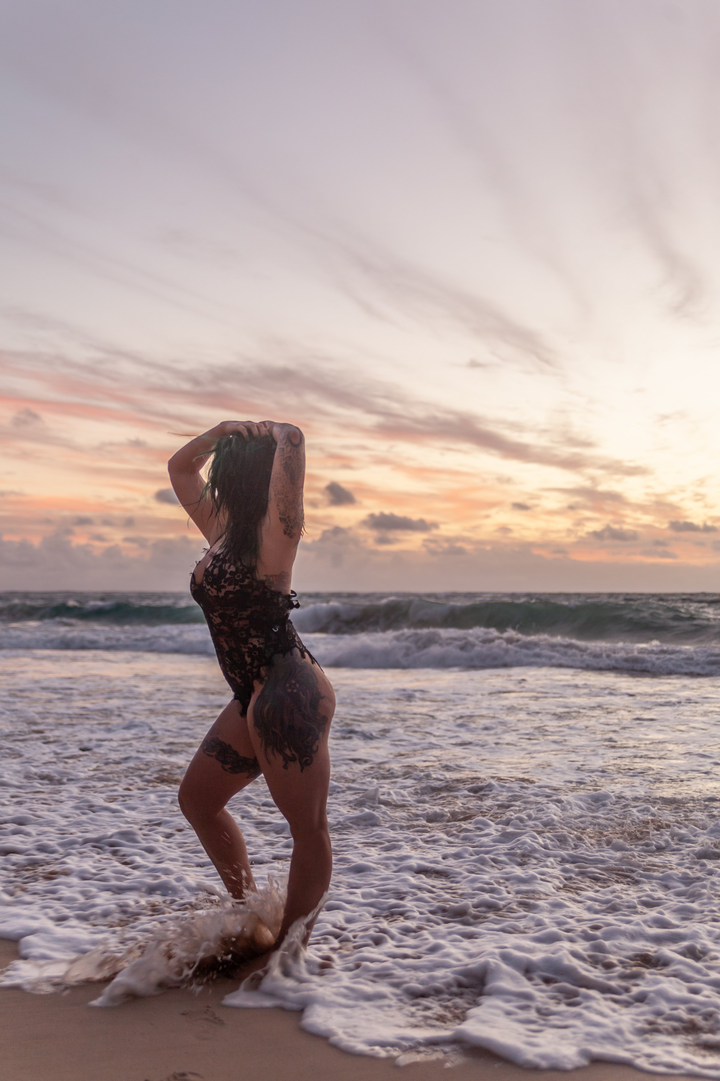 A woman with tattoos in a black lace swimsuit standing at the beach during sunset, with waves crashing around her in hawaii.