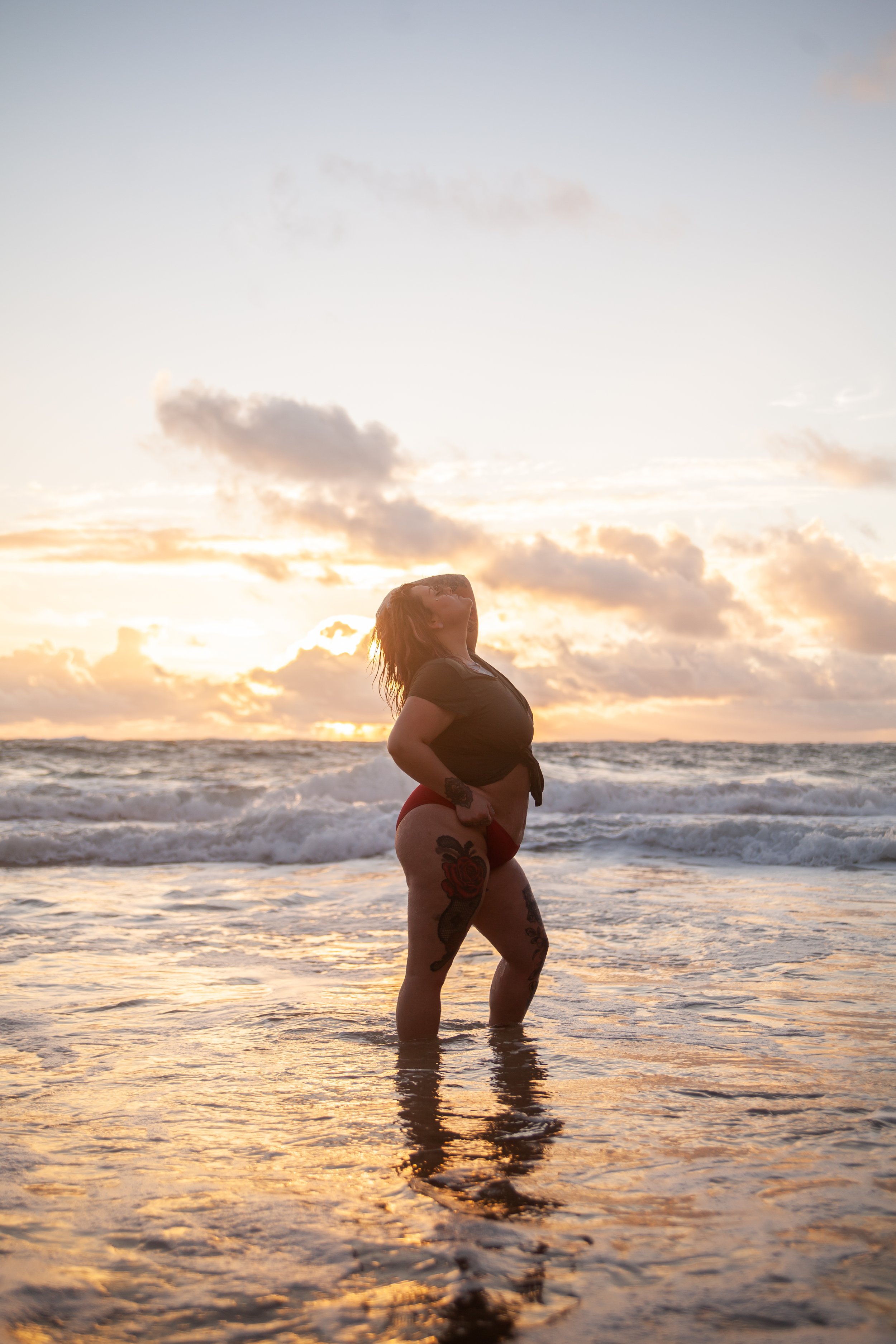 A woman with tattoos standing in the ocean at sunset, with her head tilted back and hands on her hips.