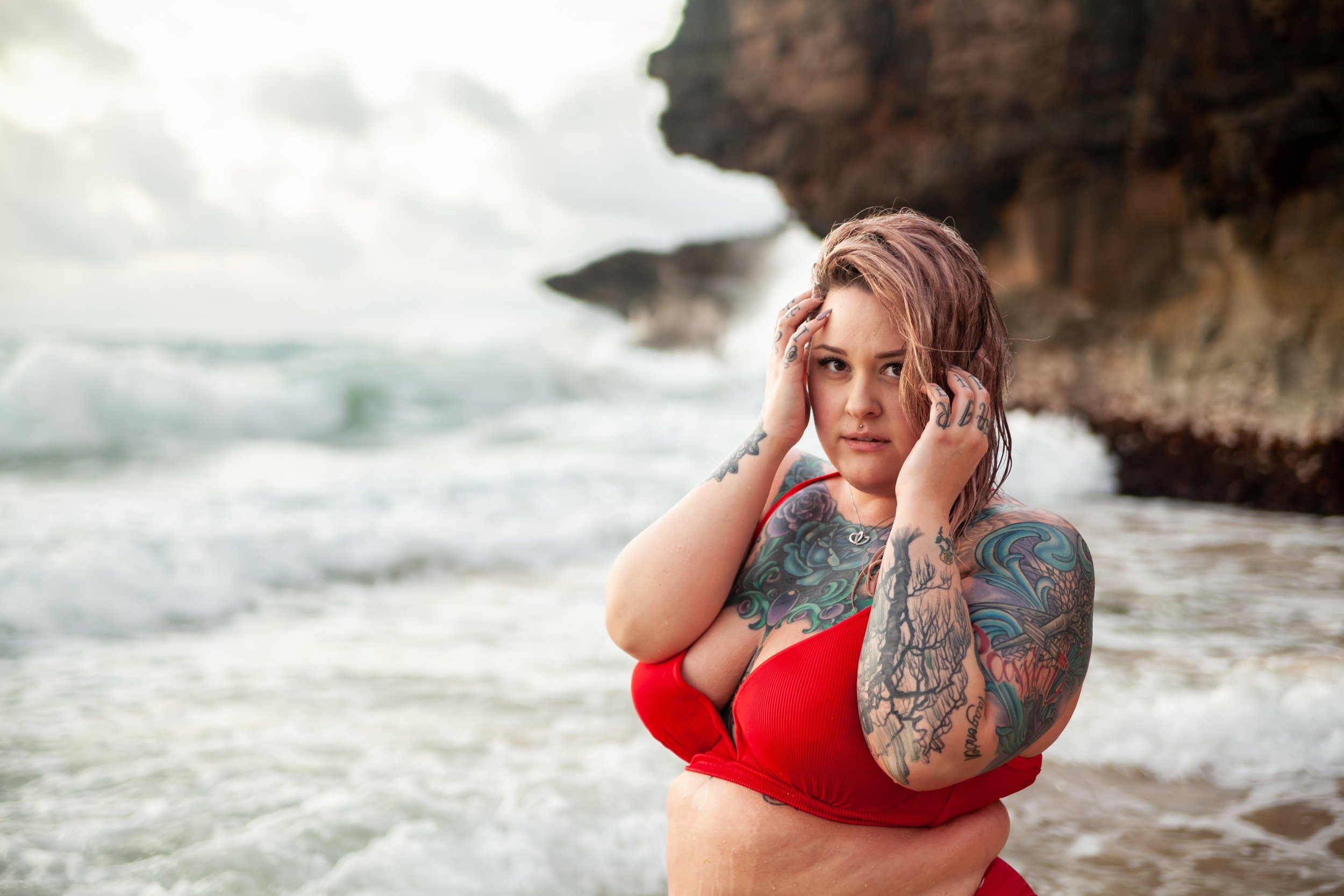 A young woman with tattoos in a red swimsuit standing in the ocean near rocky cliffs, holding her head with her hands, looking at the camera with a serious expression.