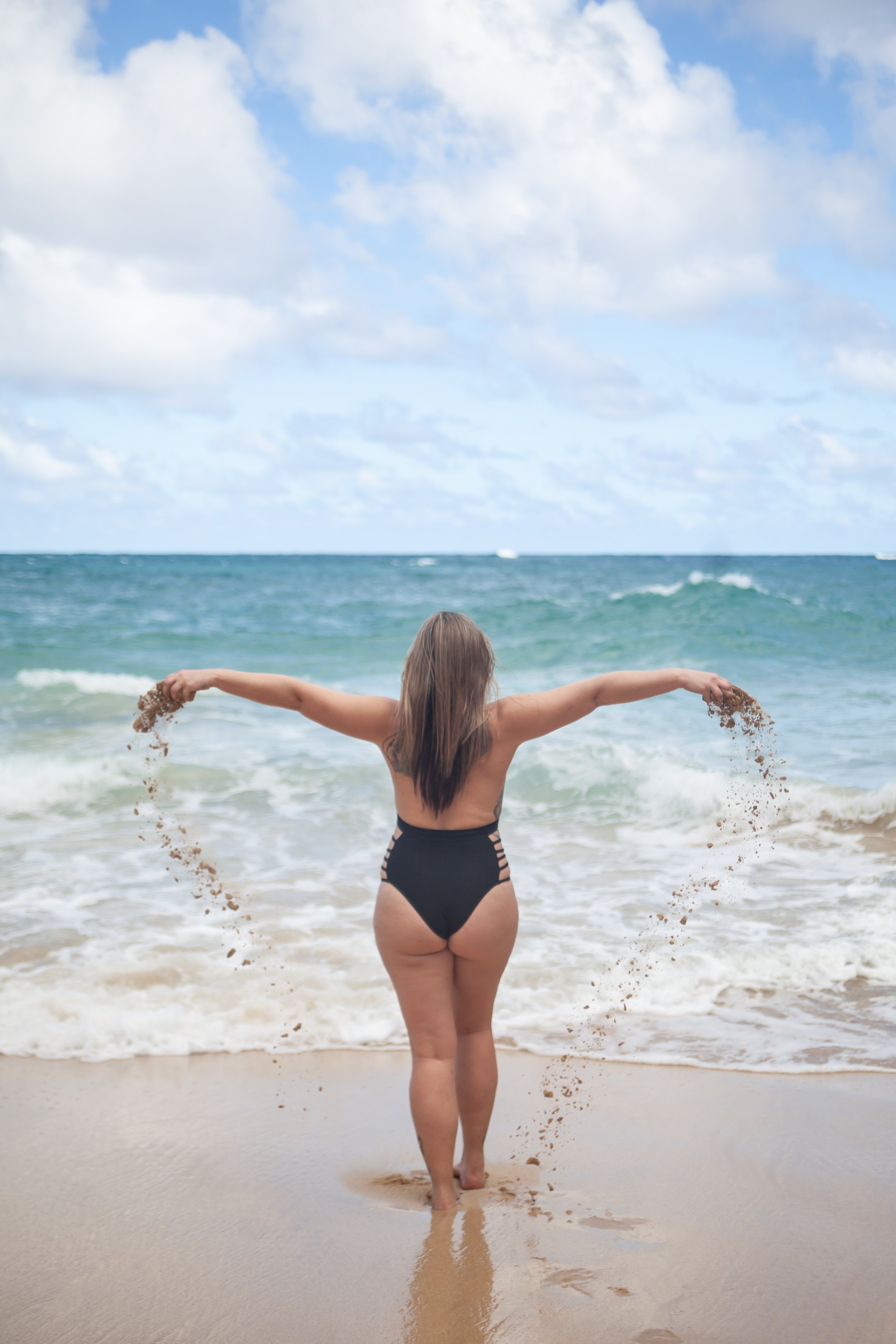 Woman in a black swimsuit walking into the ocean on a sandy beach with arms outstretched, holding sand, under a partly cloudy sky.