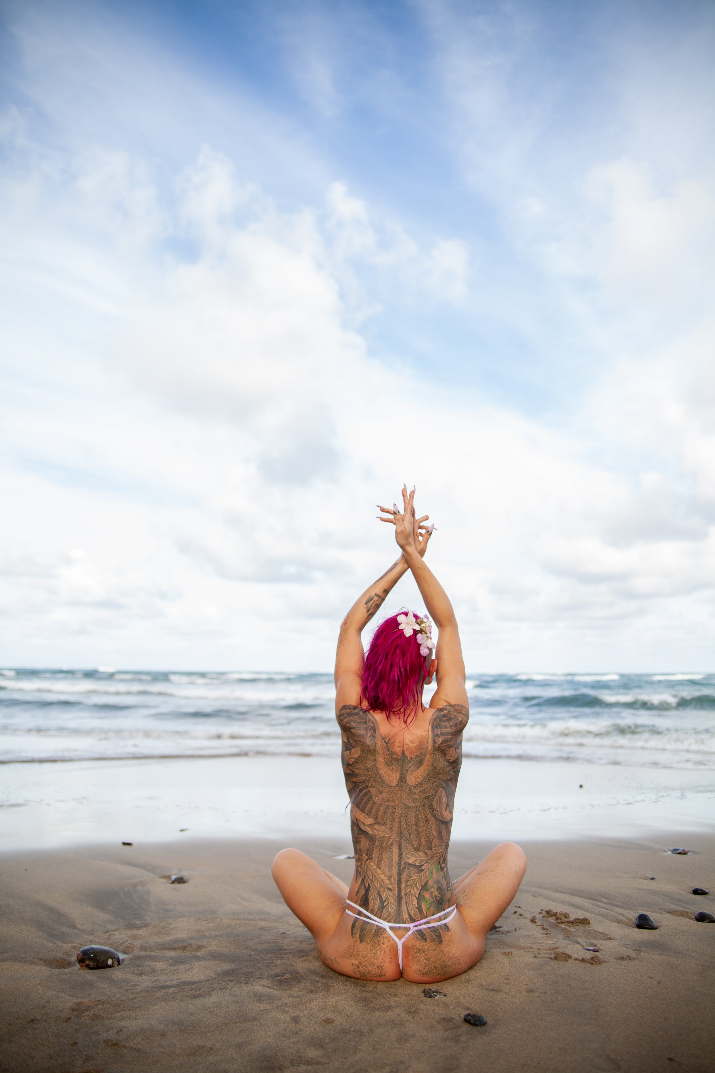A woman with pink hair and tattoos sitting on the beach in a meditative pose, facing the ocean, with hands raised above her head and crossed fingers, under a partly cloudy sky.