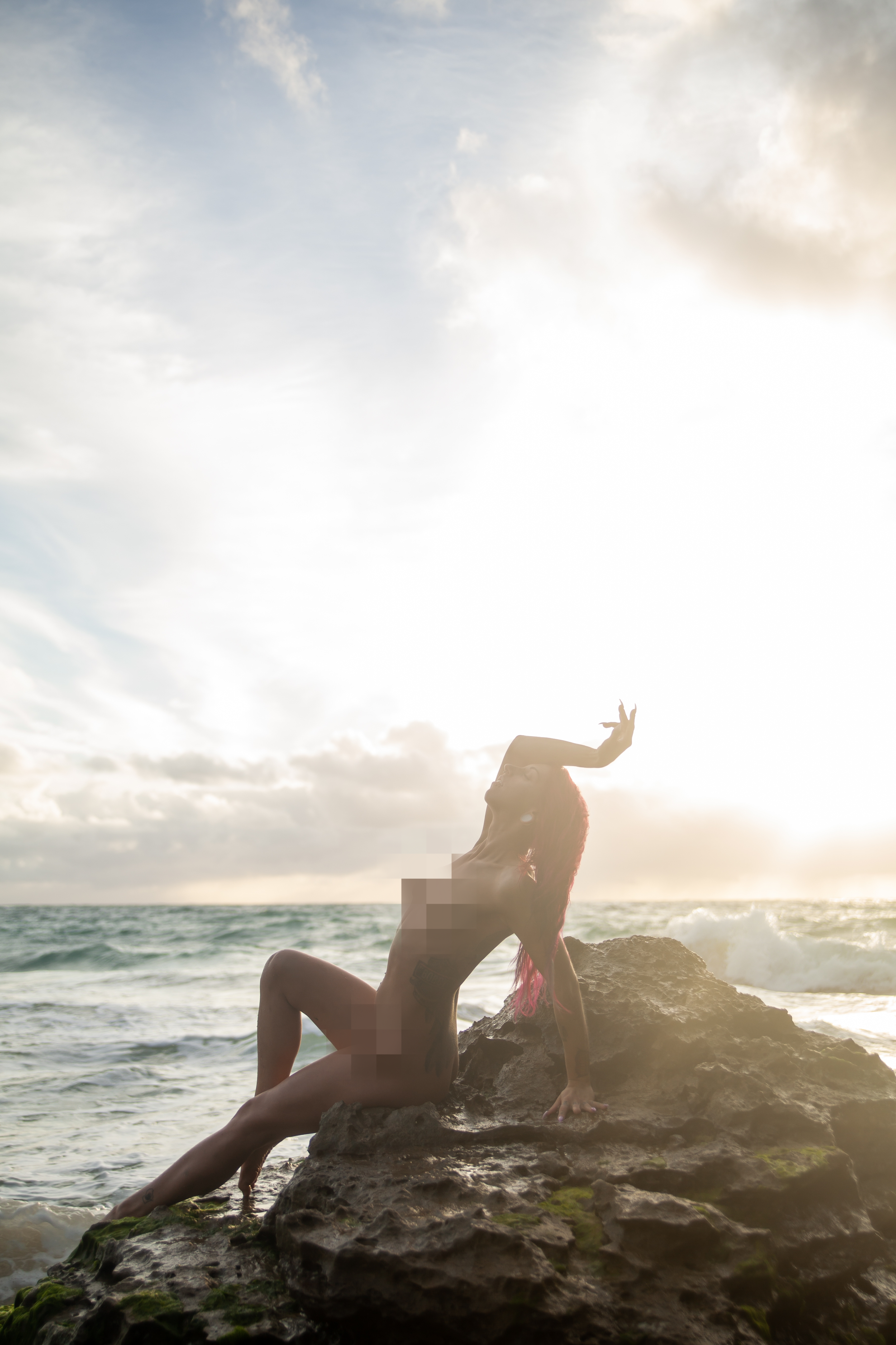 A woman in hawaii with pink hair posing nude on a rock near the ocean at sunset, with her body partially pixelated for modesty.