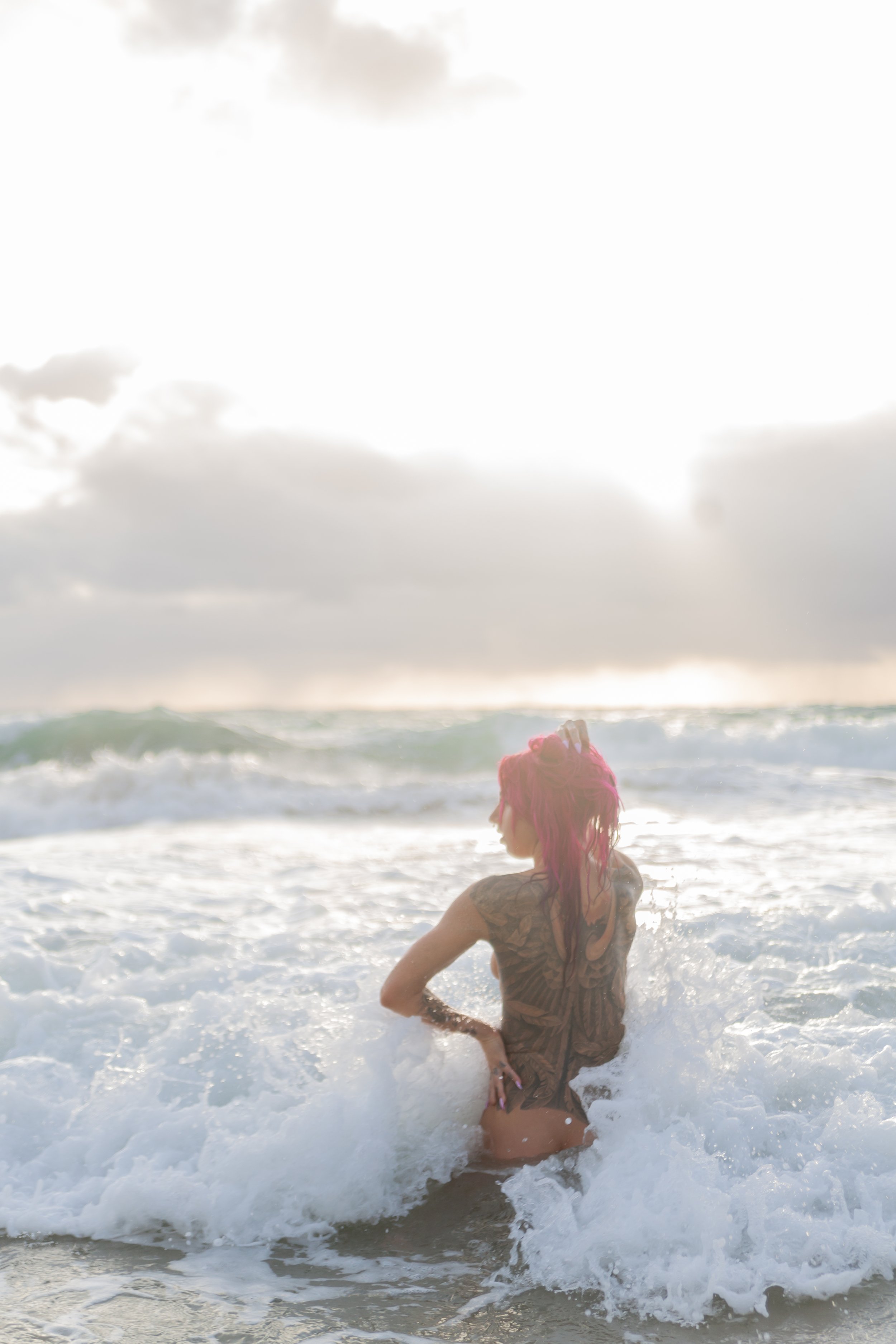 Woman with pink hair and tattoos standing in the ocean with waves crashing around her, facing away from the camera.