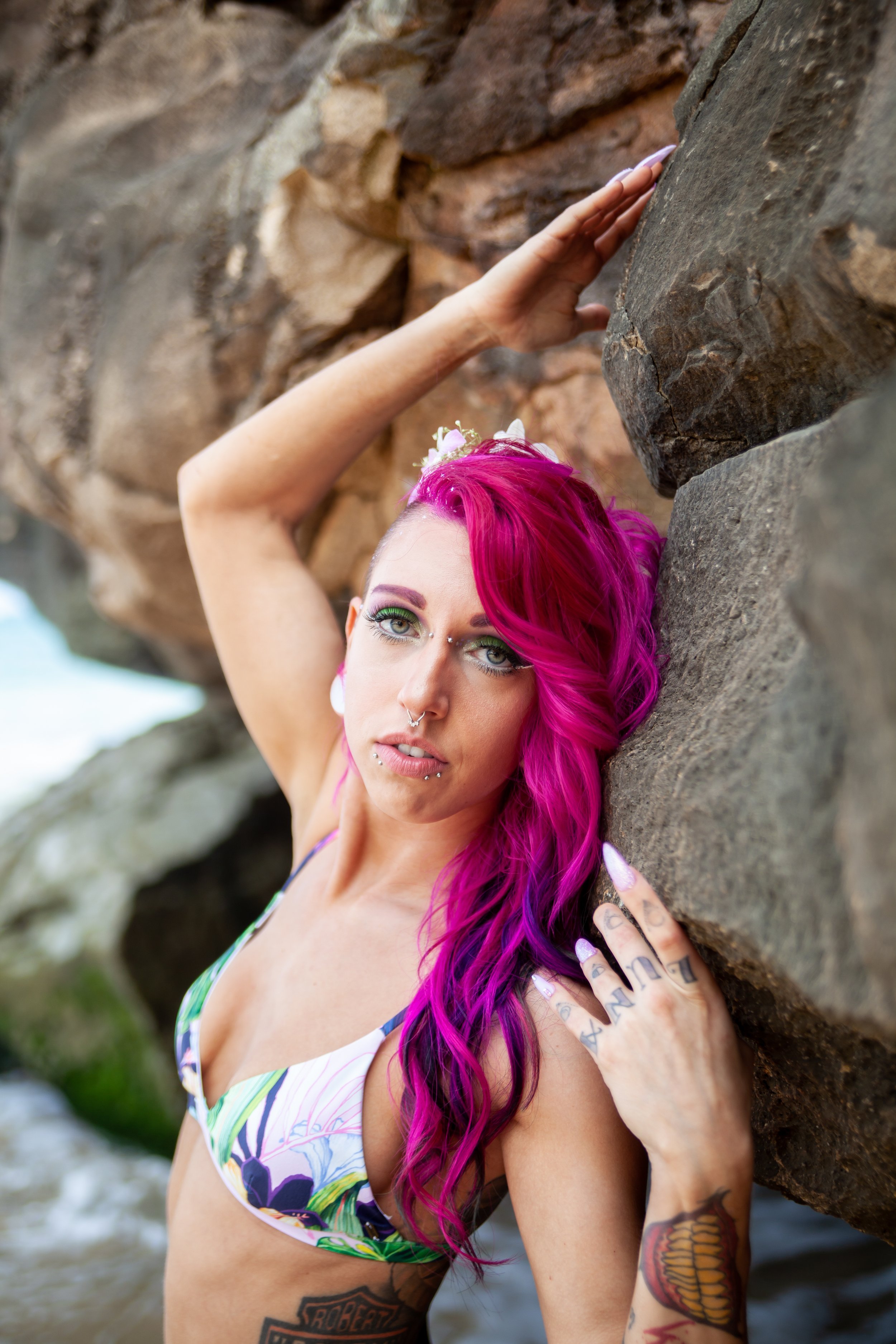woman with bright pink hair, tattoos, and piercings leaning against a large rock near the beach