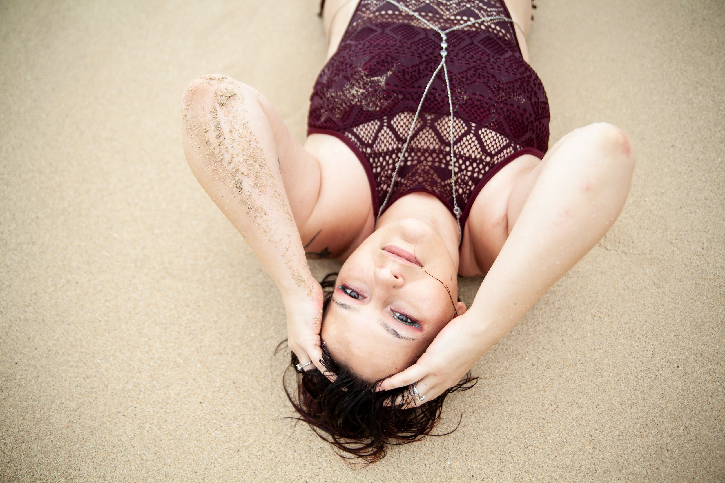 Woman lying on sandy beach looking up, wearing lace dress, with sandy arm and sand on her leg, holding her hair with hands.