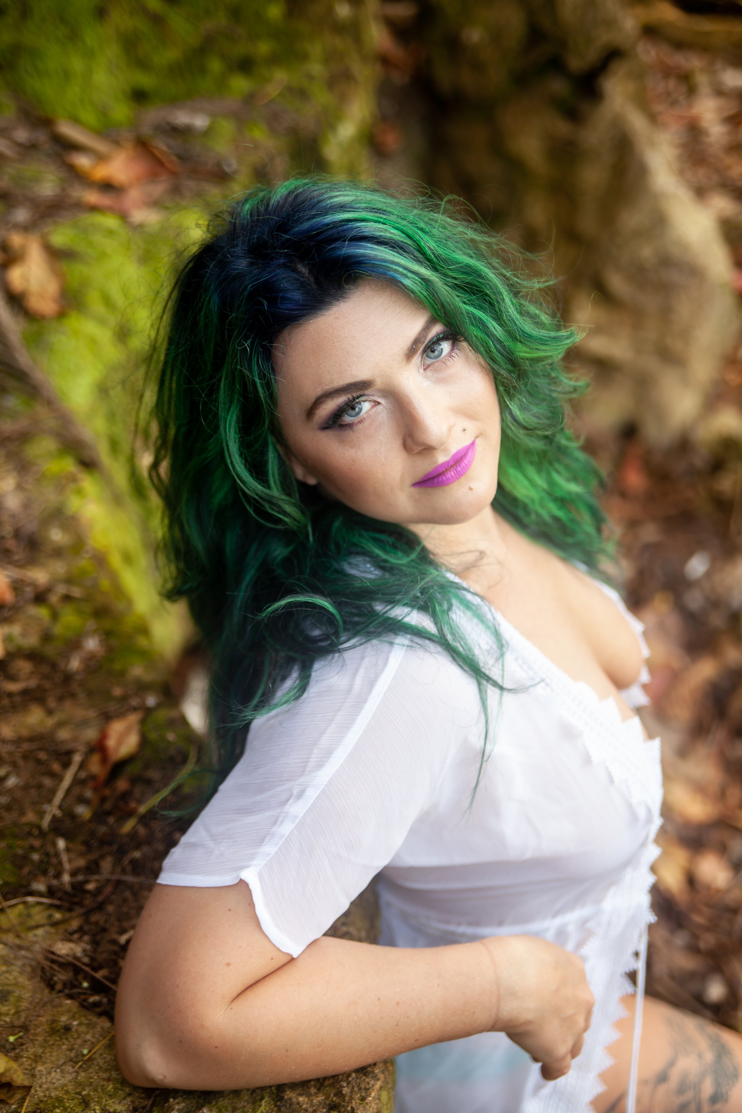 A woman with vibrant green and blue hair, wearing a white blouse, looks up at the camera in a natural outdoor setting with moss-covered rocks and fallen leaves.