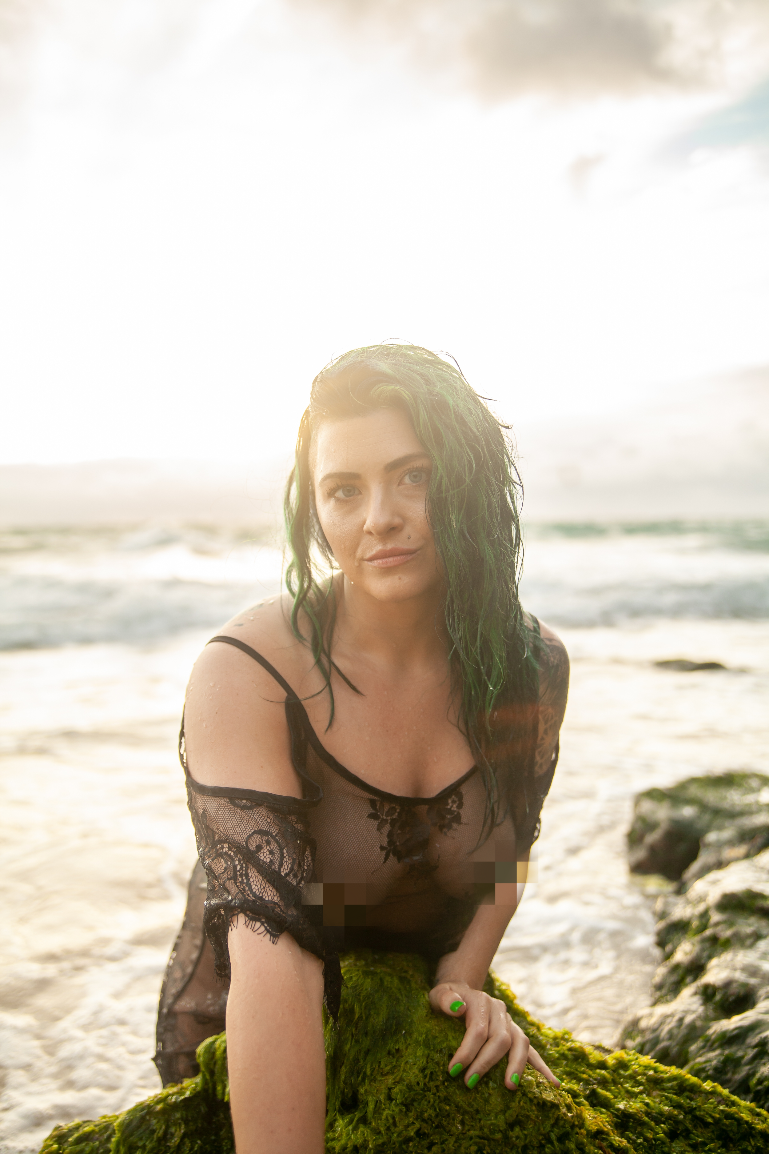 Woman with green-highlighted black hair and tattoos leaning on a moss-covered rock at the beach during sunset.