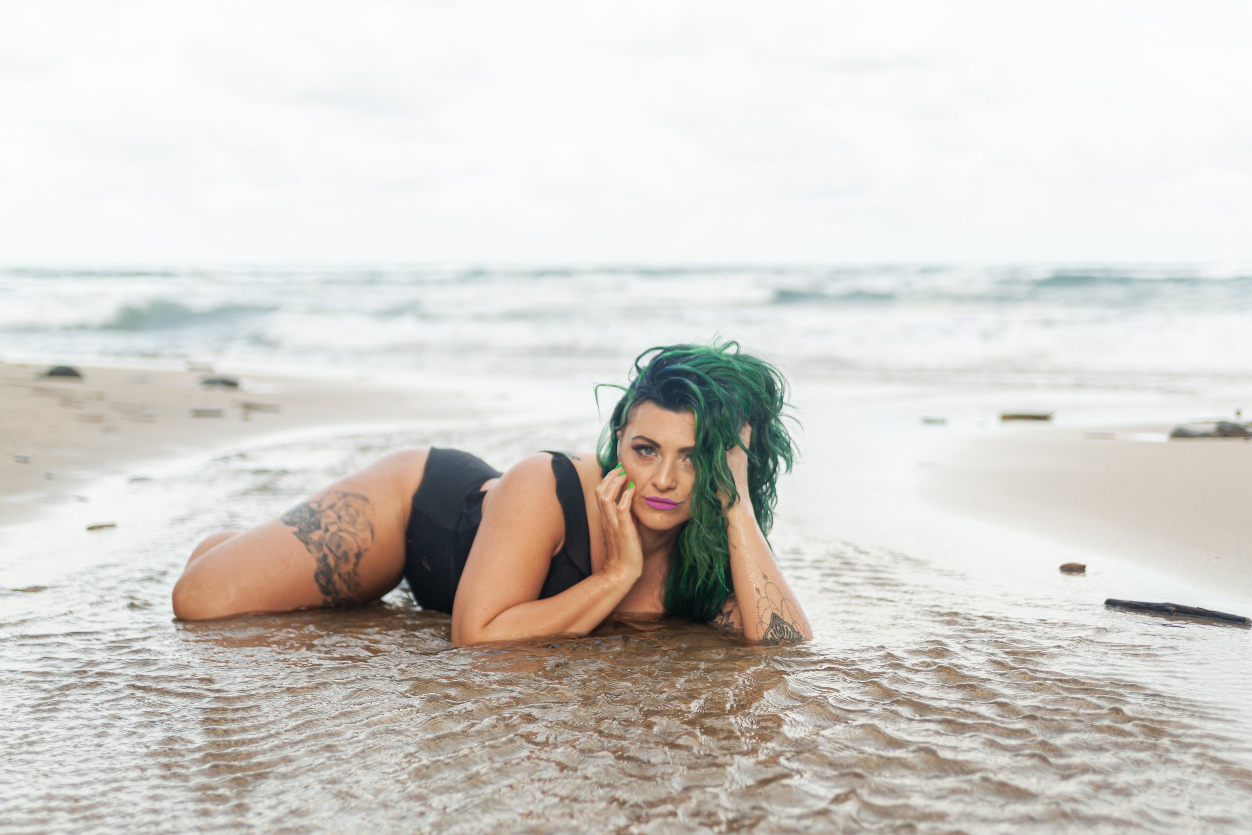 Woman with green hair lying on a sandy beach in shallow water, looking at the camera.