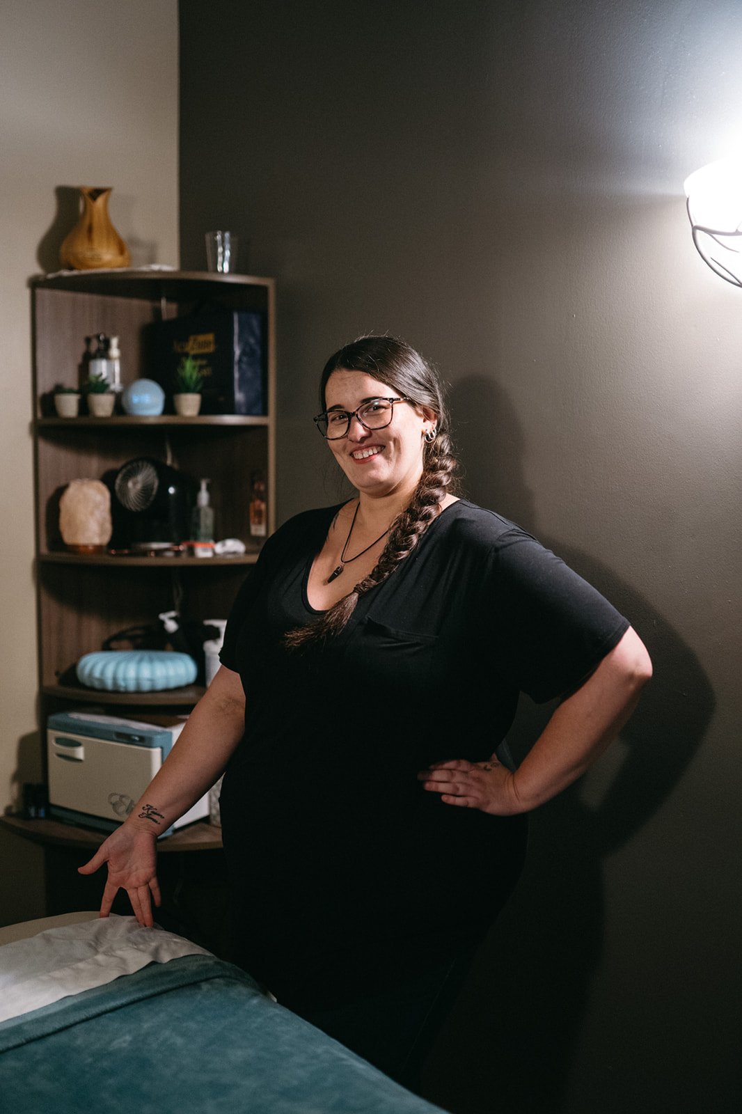 Ashleigh, Massage Therapist at Hometown Chiropractic & Wellness in Nolensville, TN, smiling woman with glasses and a braid, wearing a black shirt, standing next to a massage table with dark gray walls and a wooden shelf.
