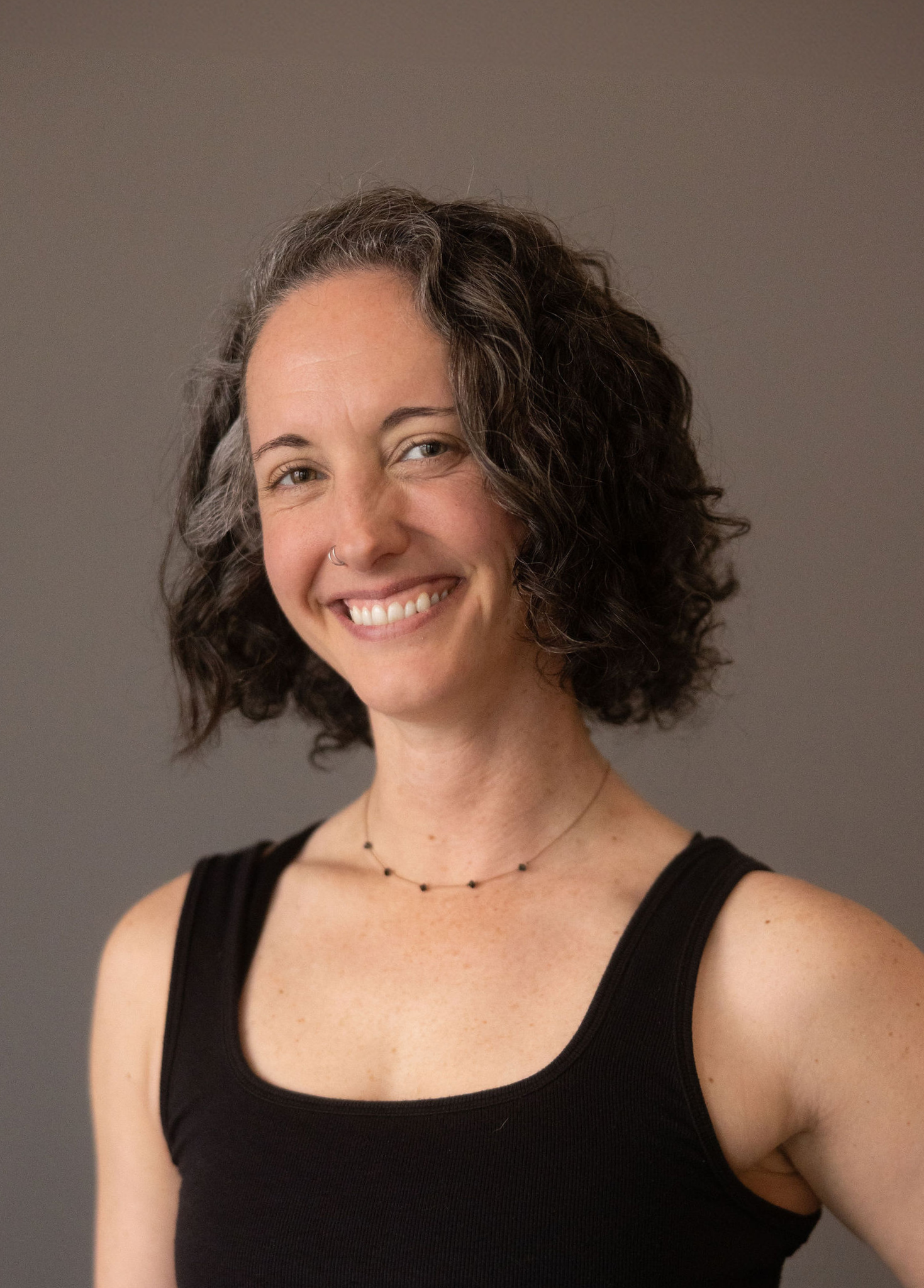 A woman with curly dark hair wearing a black tank top and a delicate black necklace, smiling against a plain gray background.