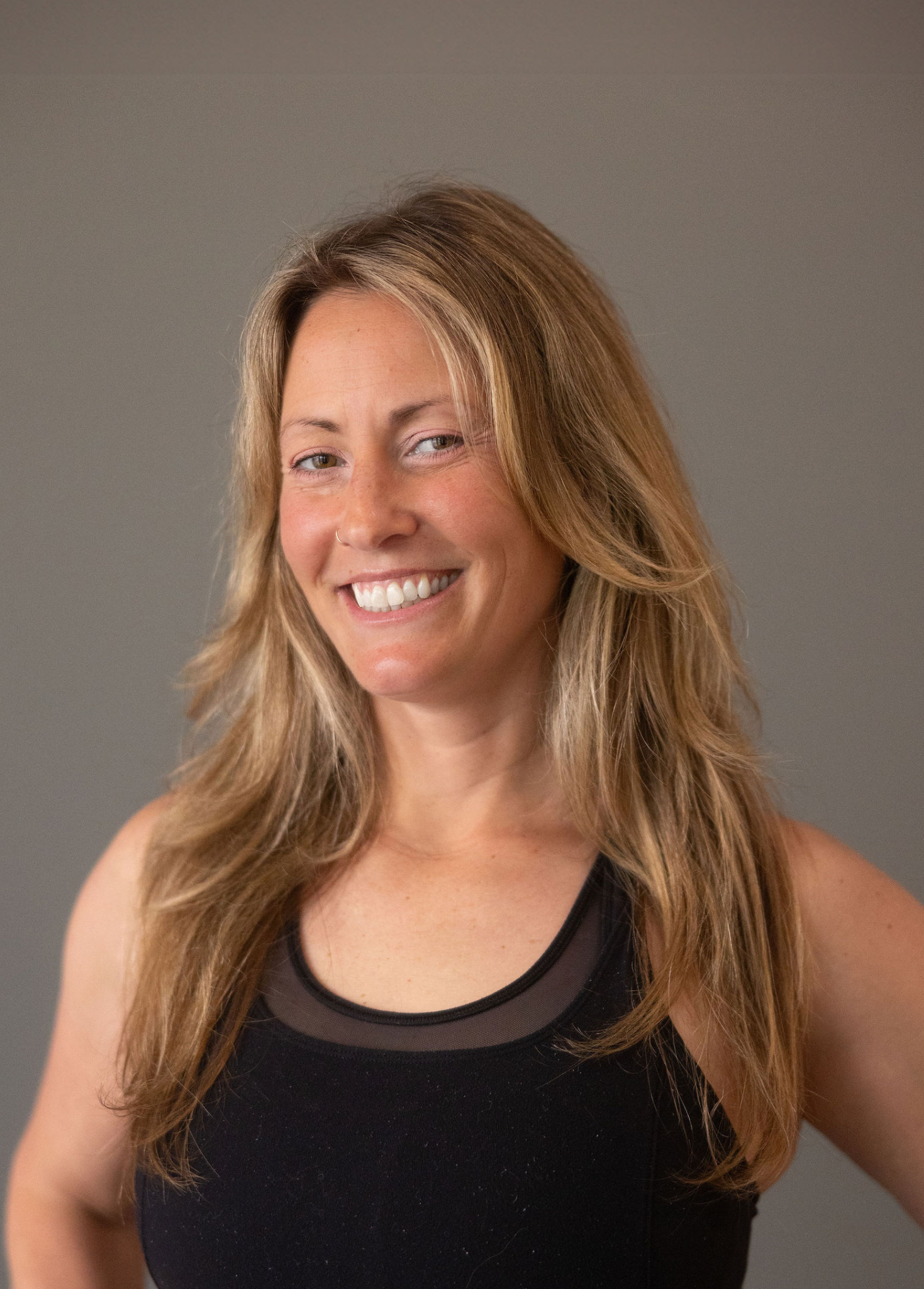 Close-up of a smiling woman with long, wavy blonde hair, wearing a black sleeveless top, against a plain gray background.