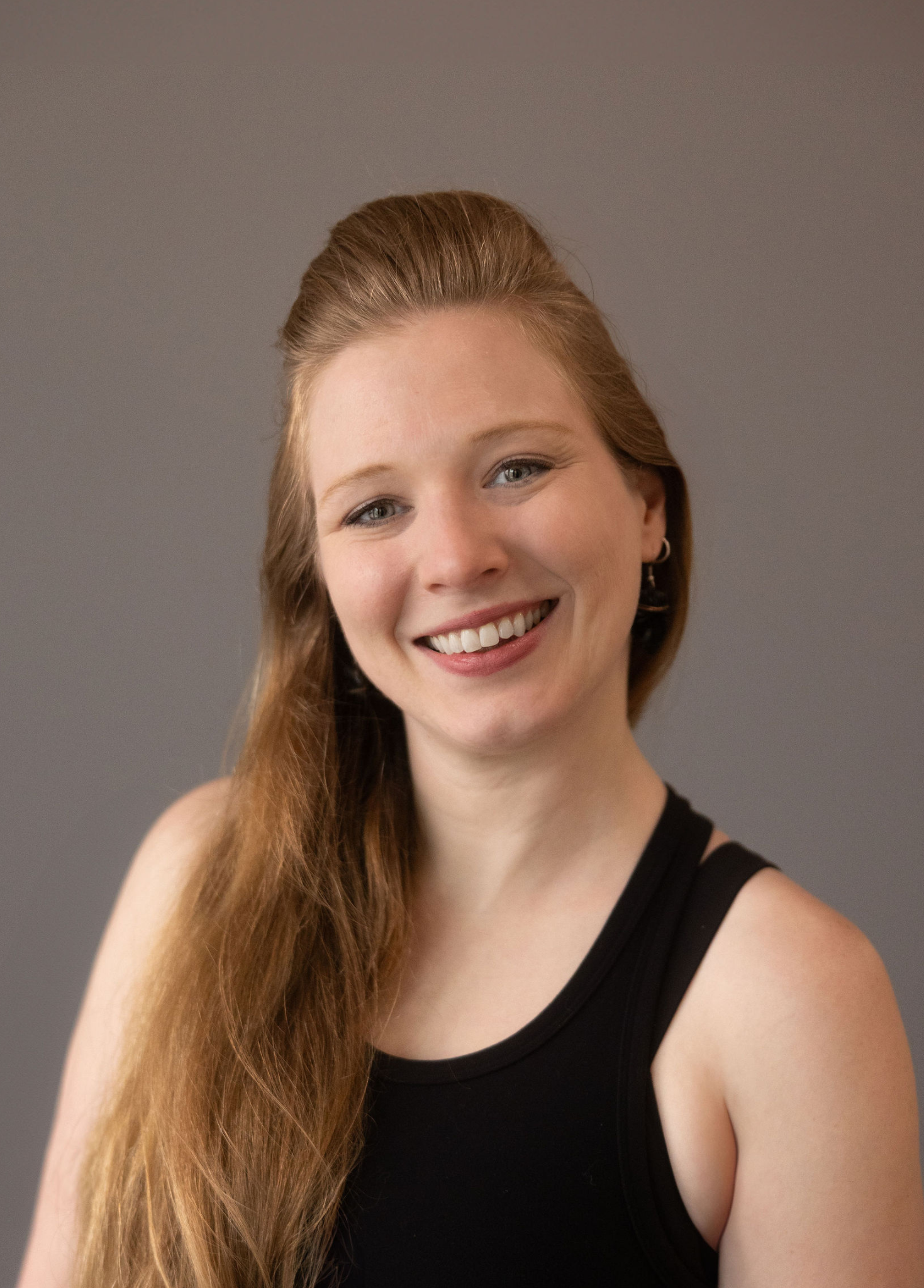 Close-up of a woman with long, reddish-brown hair, smiling and wearing a black sleeveless top, against a plain gray background.