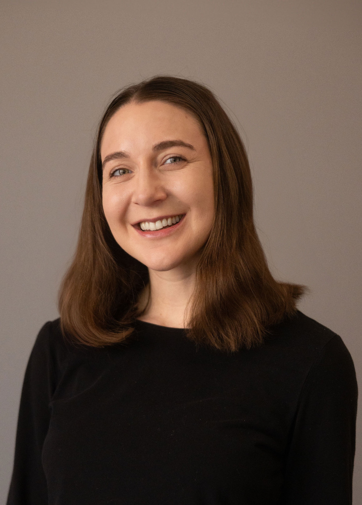 A woman with long brown hair wearing a black shirt smiling at the camera, against a neutral background.