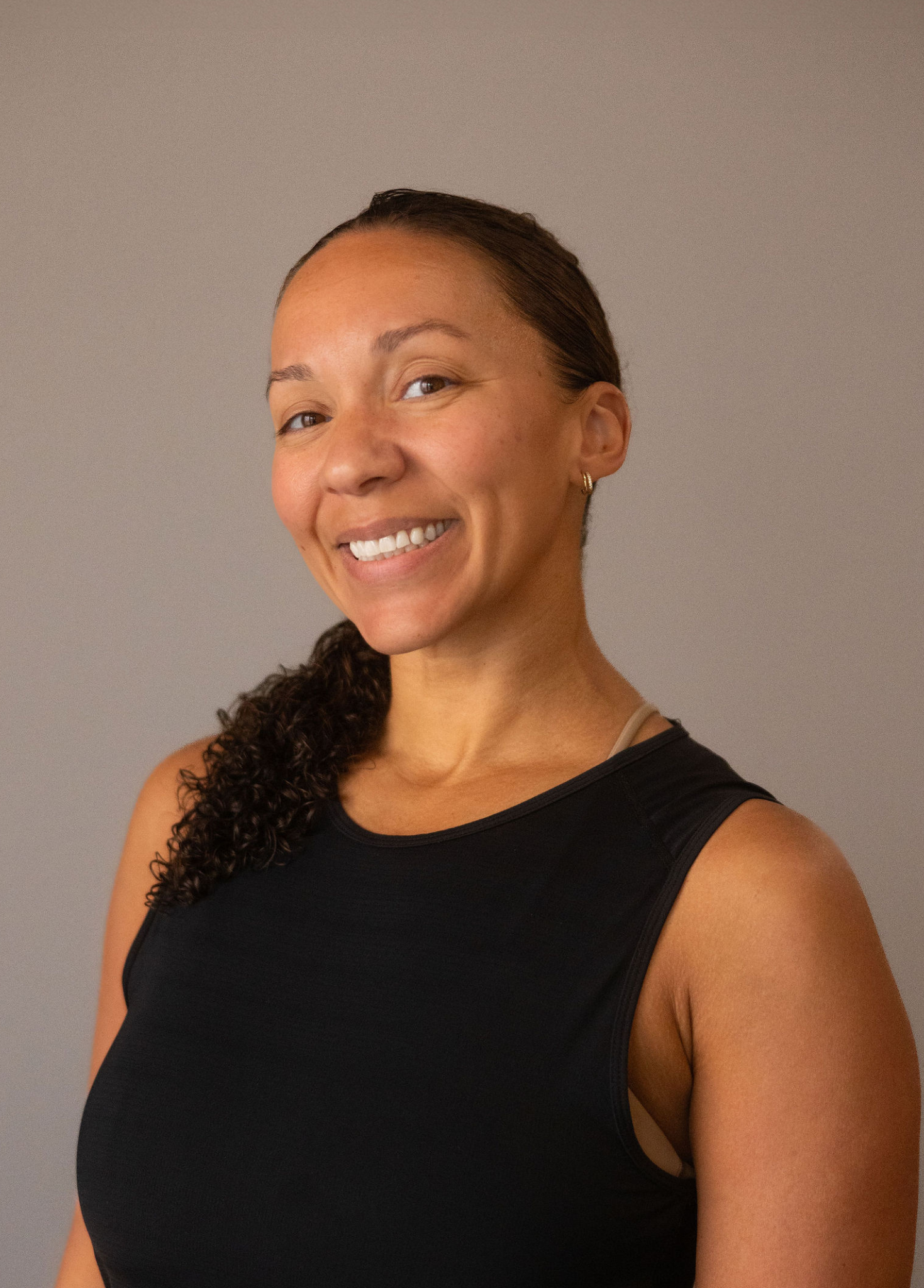A woman with brown hair tied back, smiling, wearing a black athletic tank top, standing against a plain gray background.