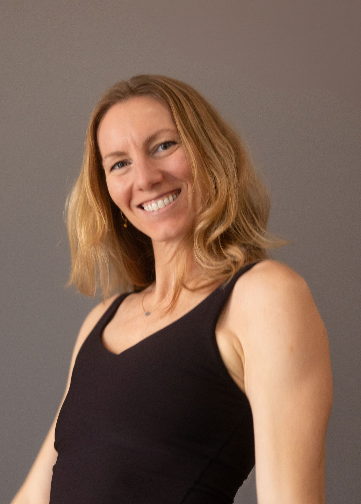 A smiling woman with wavy, shoulder-length blonde hair, wearing a black sleeveless top, standing against a plain gray background.