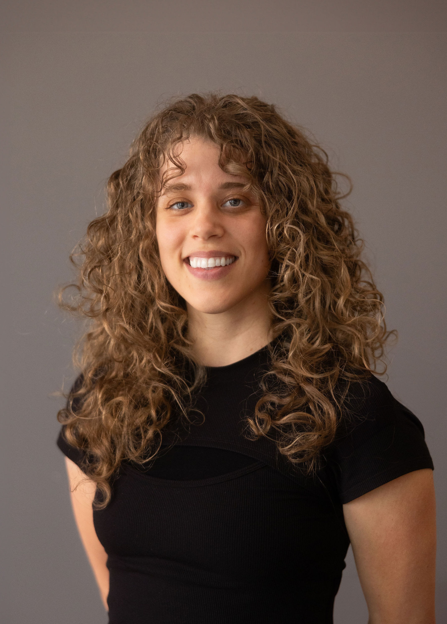 Portrait of a young woman with curly light brown hair and blue eyes, smiling, wearing a black top, against a gray background.