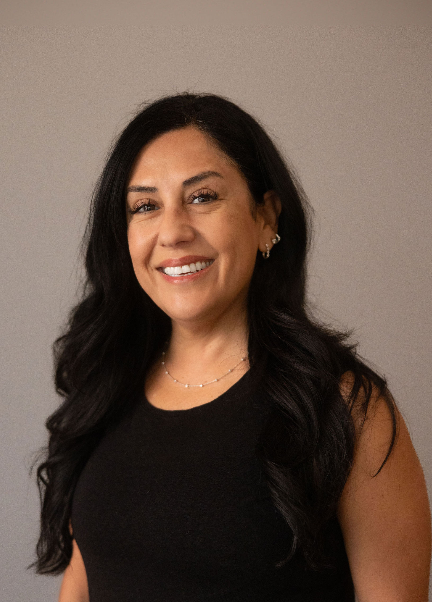 Close-up of a smiling woman with long dark hair wearing a black sleeveless top, silver jewelry, and a neutral background.