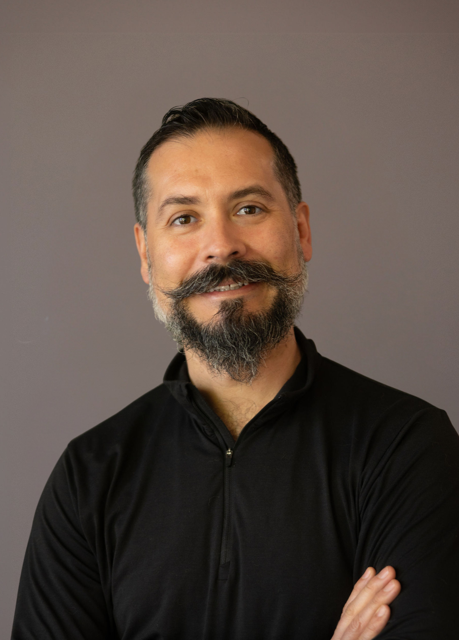 A man with a beard and mustache, wearing a black zip-up jacket, smiling slightly with arms crossed, against a plain gray background.