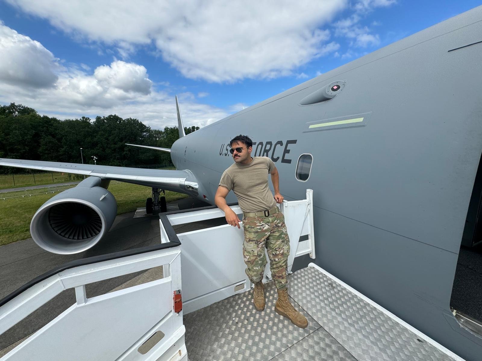A man in military camouflage pants, tan t-shirt, and tan boots leaning against the side of a large U.S. Air Force cargo plane, with a grassy airfield and trees in the background under a partly cloudy sky.