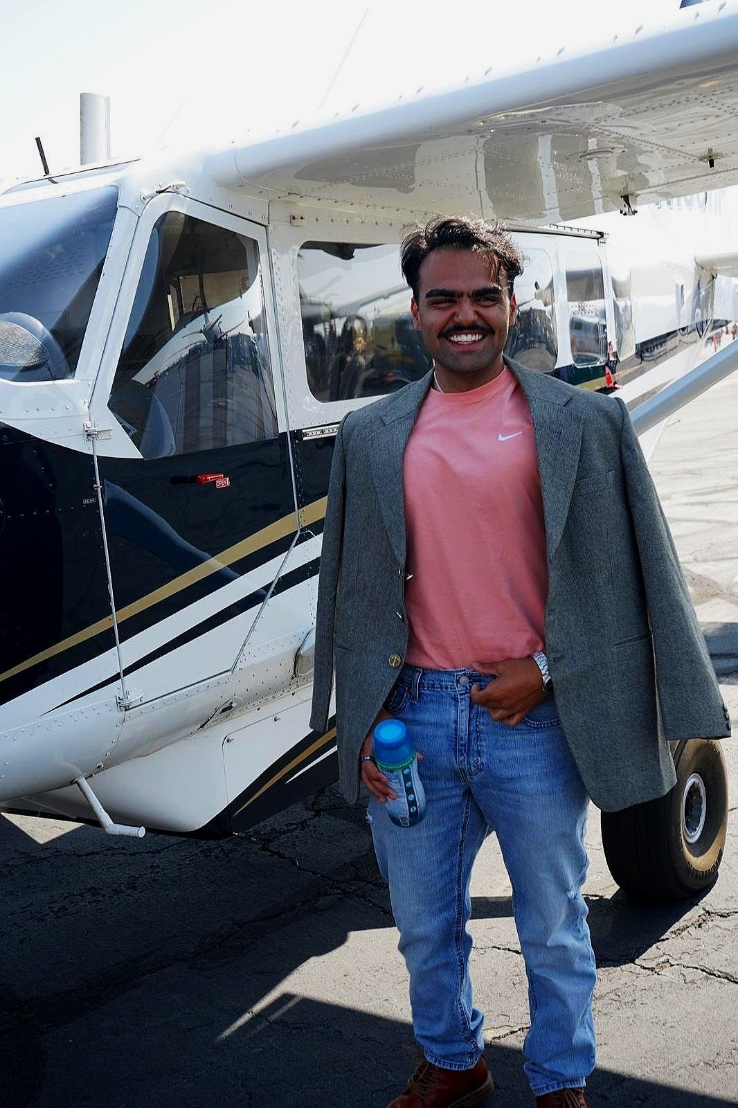 A man smiling and holding a water bottle standing in front of a small aircraft on a tarmac at an airport.