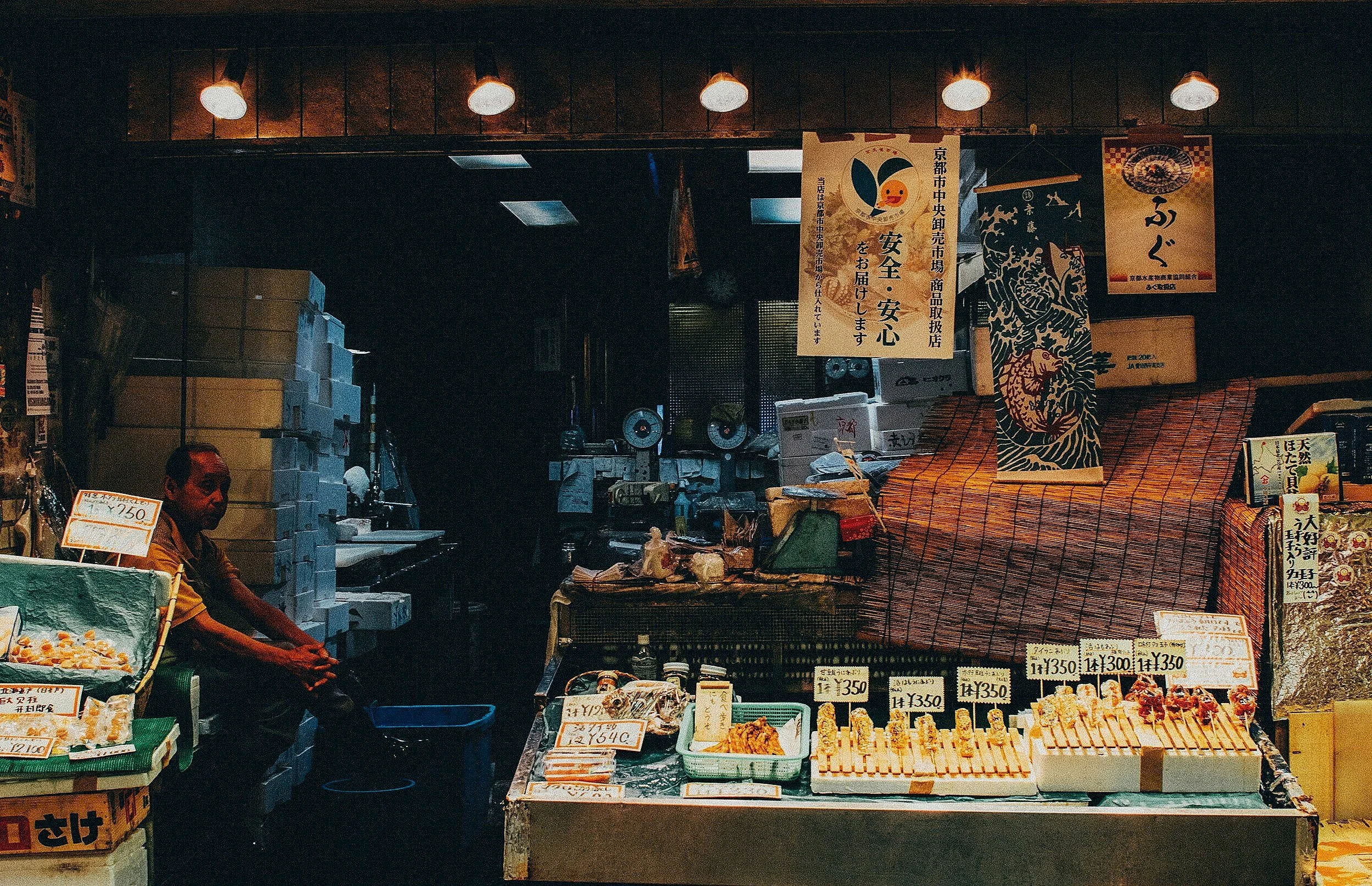 A Japanese market stall sells seafood with a man sitting behind the counter, signs displaying prices, and various packaged fish products on display.