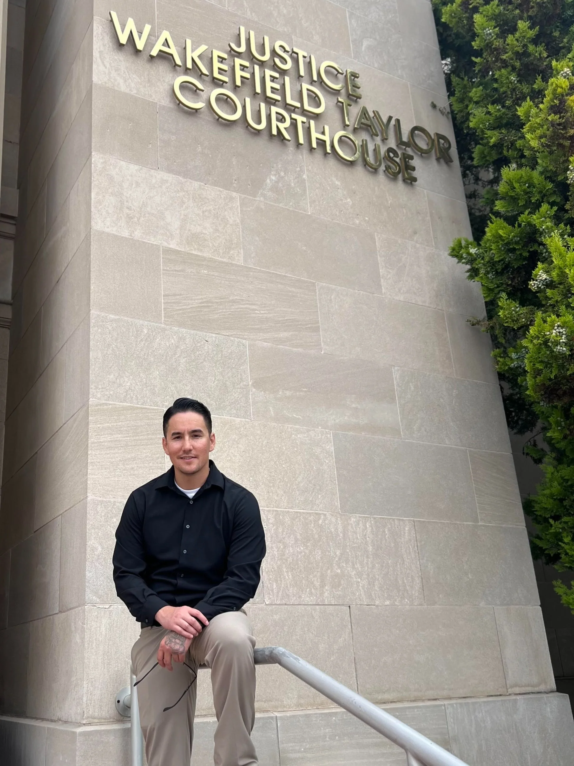 Person sitting on a railing in front of the Justice Wakefield Taylor Courthouse, wearing a black shirt and beige pants, with trees partially visible.
