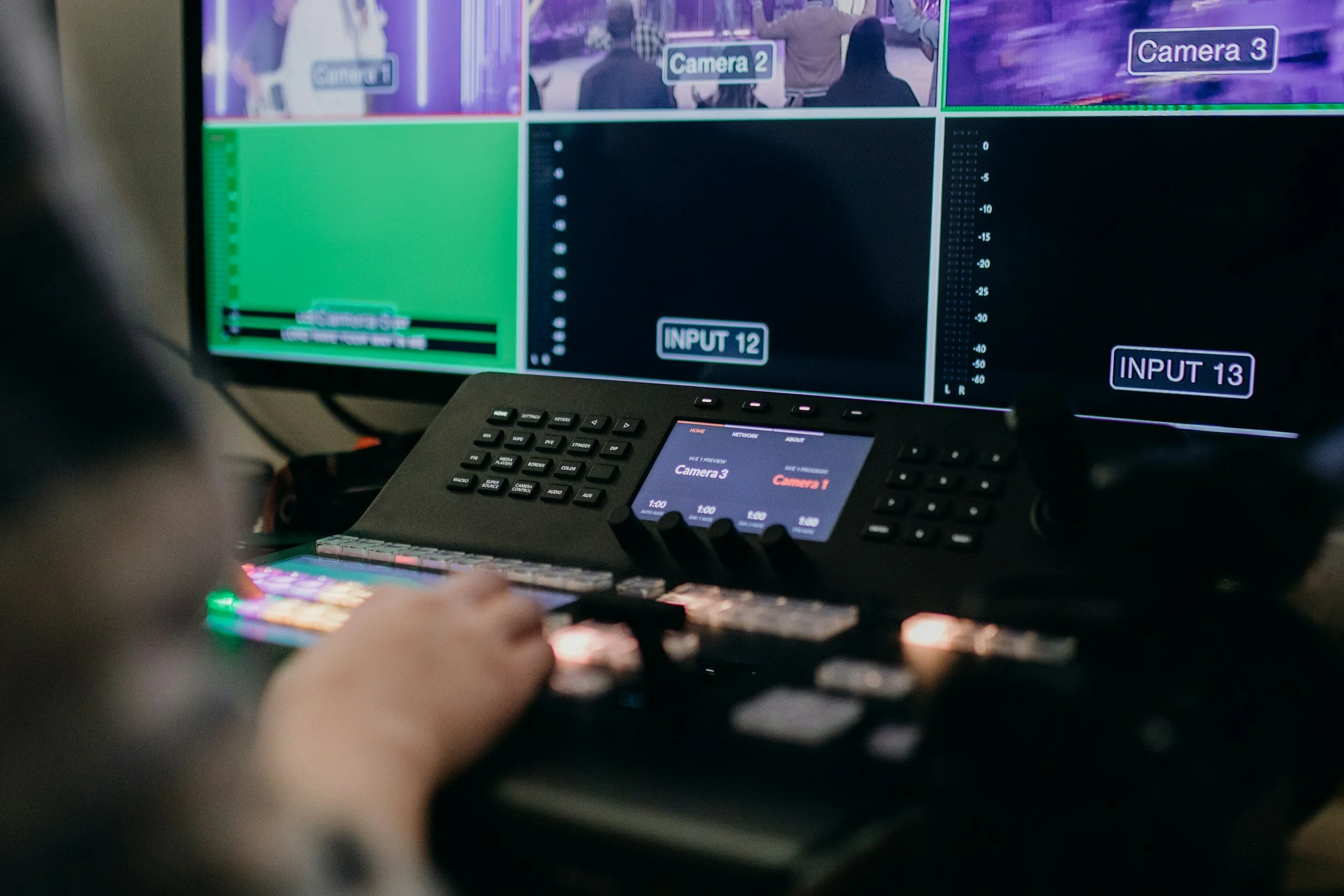 Close-up of video control and monitoring equipment with a person operating a control panel in front of a large screen displaying multiple camera feeds labeled Camera 1, Camera 2, and Camera 3.