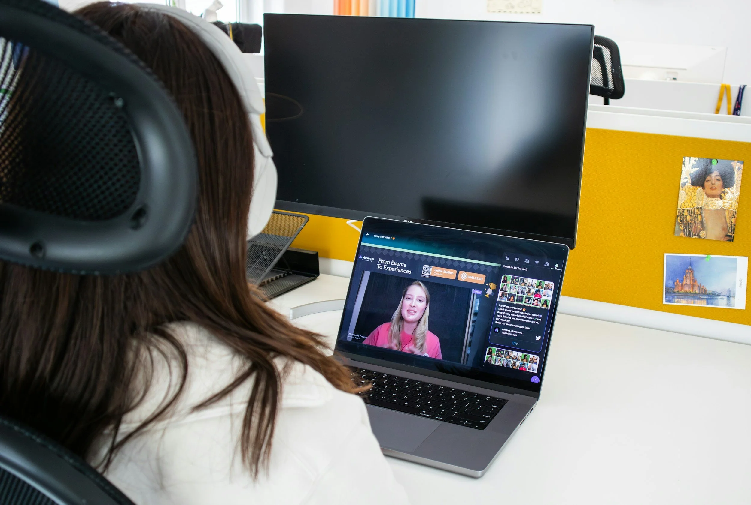 A woman wears headphones and looks at a laptop computer with a video conference or presentation visible on the screen, in an office setting with a yellow partition and photos on the wall.