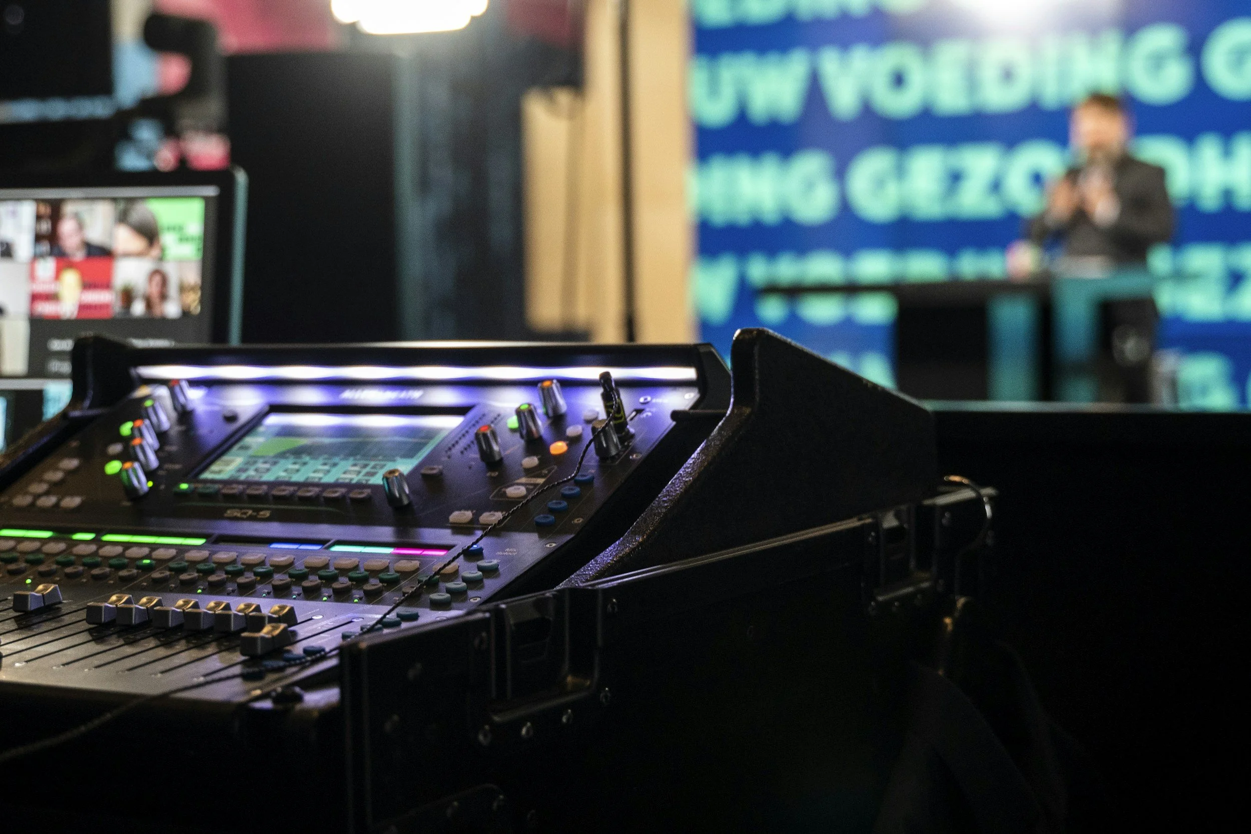 Close-up of a professional sound or lighting control mixing console on a stage, with a blurred presenter or speaker in the background on a blue screen.