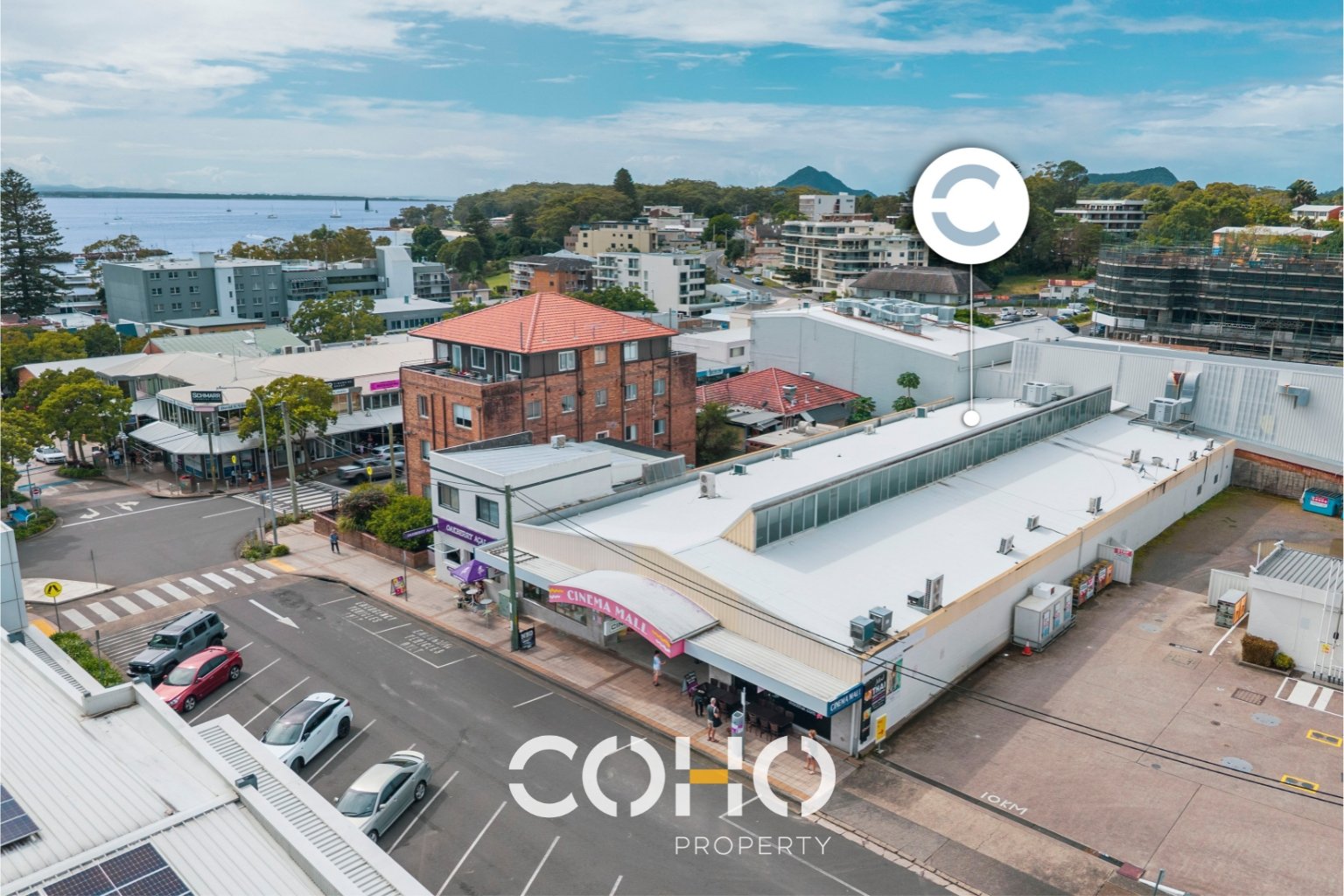 Aerial view of a shopping mall named 'CINEMA MALL' with parking lot, surrounding buildings, and a waterfront in the distance under a partly cloudy sky.