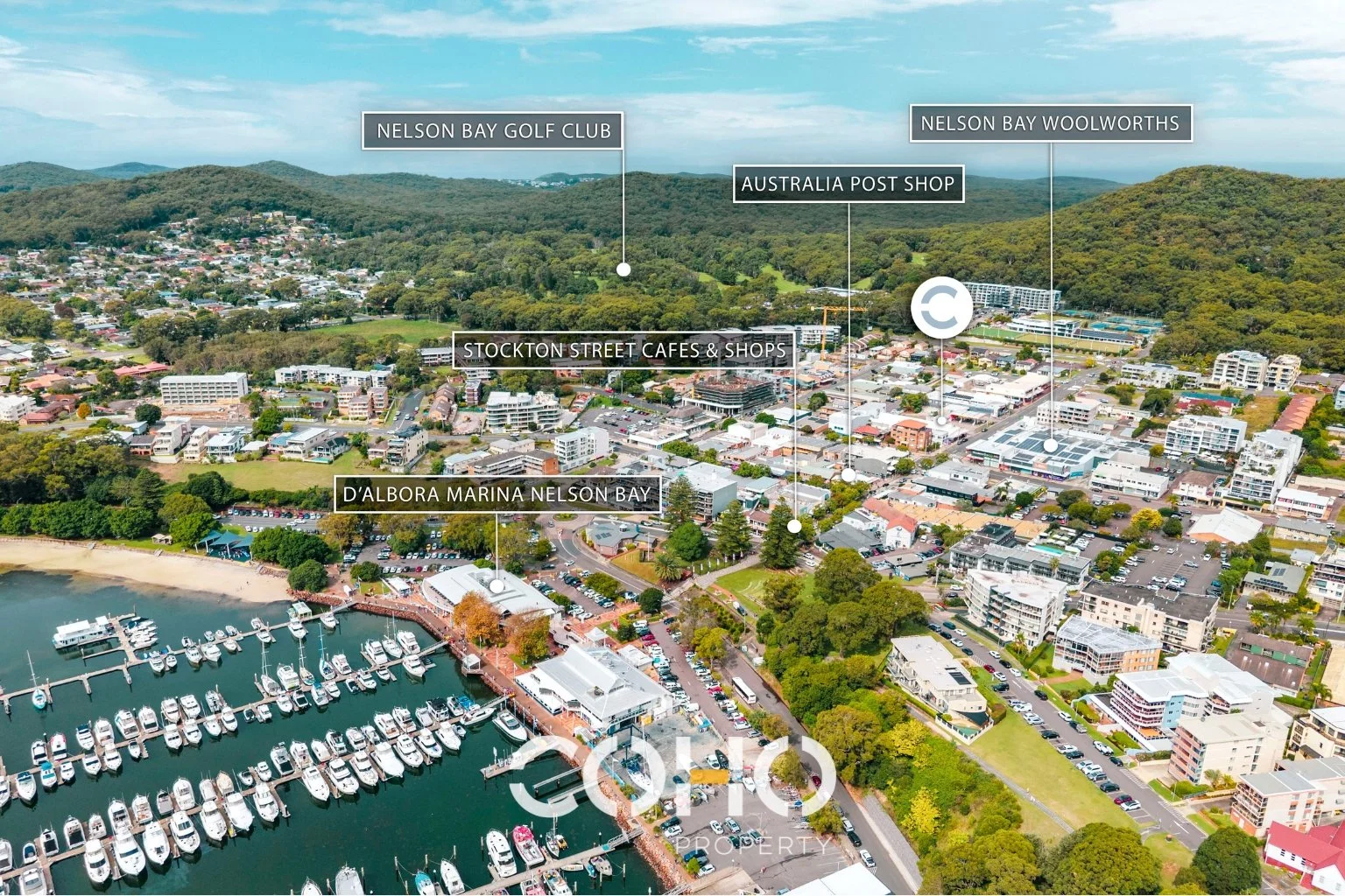 Aerial view of Nelson Bay, showing Marina area with boats, shopping and dining district including Stockton Street cafes and shops, nearby golf courses, and green hilly landscape in the background.
